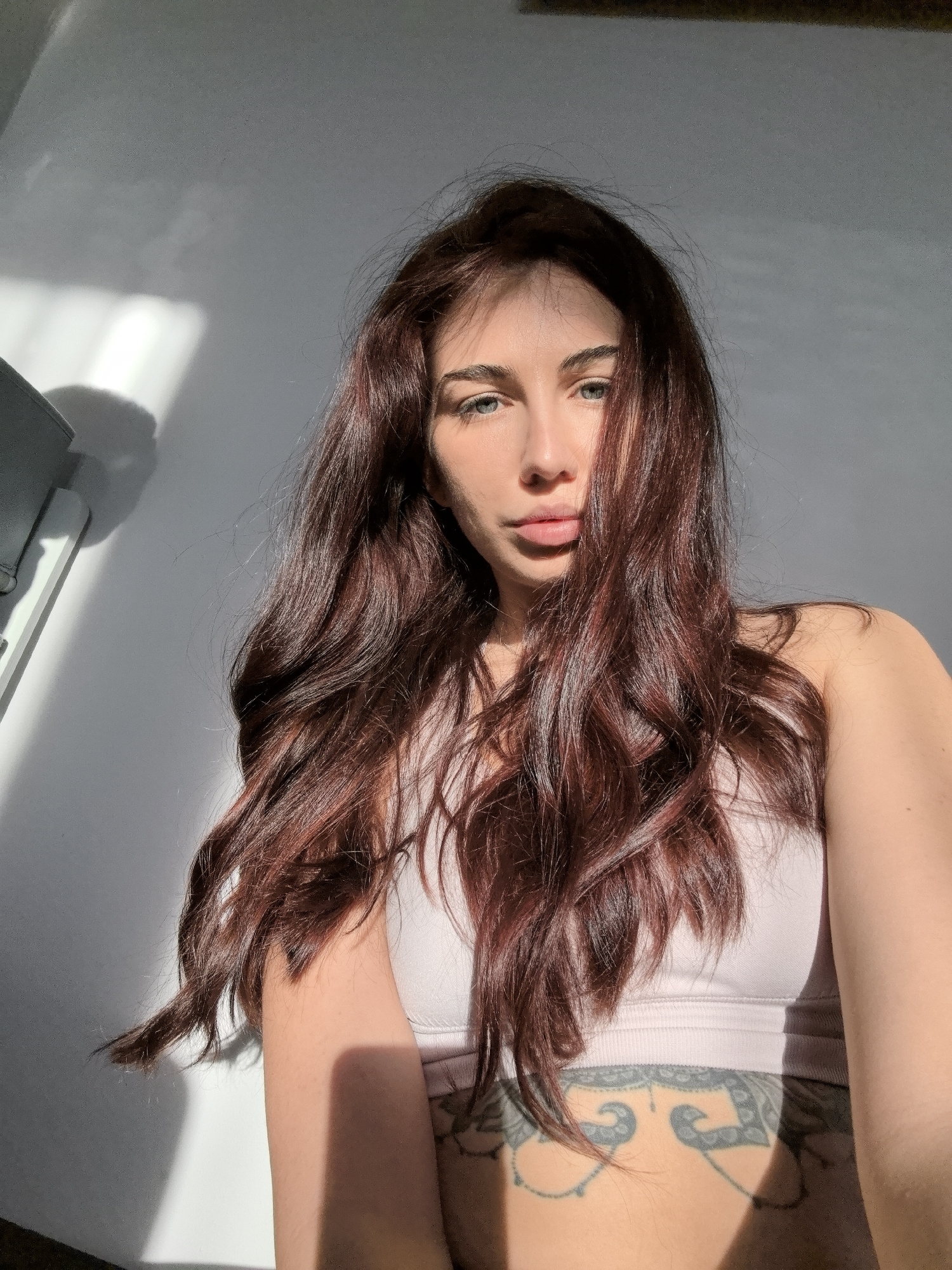 A woman with long, auburn hair and light eyes poses indoors in a casual, low-angle shot. She wears a light pink sports bra, and a visible tattoo adorns her lower abdomen. Sunlight creates stark shadows on her face and the adjacent wall, contrasting with the overall brightness of the scene. The background is a plain, light-colored wall with a partially visible object on the left. There is no discernible information to indicate that the image was taken in Stara Zagora, Bulgaria.