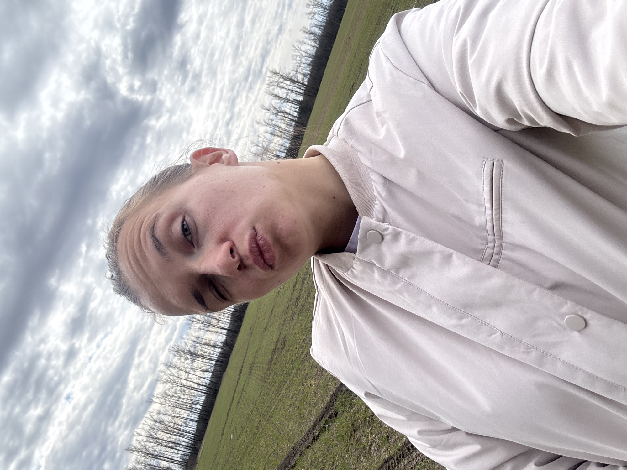 A woman is taking a selfie in a field near Razdol'e, Russia. The sky is overcast with thick clouds. A line of bare trees stands in the distance between the field and the sky. The woman is wearing a light-colored jacket. She is looking at the camera with her lips pursed. The field appears to be green with patches of brown earth.