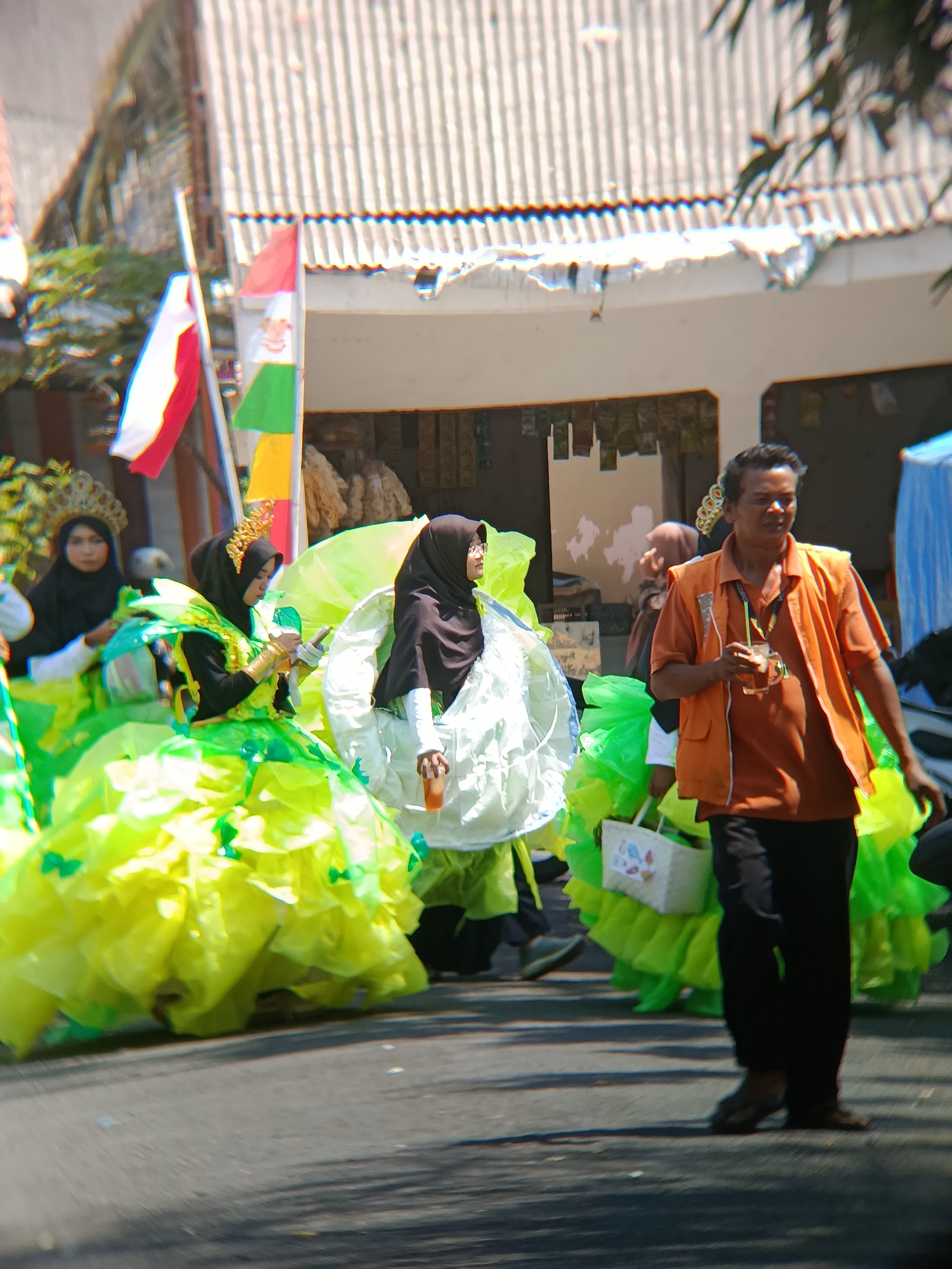 The image depicts a street scene in Desa Kebon Manis, Kecamatan Cilacap Utara, Indonesia, likely during a cultural event or parade. In the foreground, a man wearing an orange polo shirt and a vest walks while holding a cup with a straw. To his left, several individuals are adorned in elaborate, brightly colored costumes, primarily in shades of green and yellow, with one person wearing a black hijab and a golden headdress. These individuals appear to be participating in a procession. In the background, there are buildings with corrugated roofs and some foliage. Flags are visible, including one that appears to be a red and white striped flag. The overall atmosphere suggests a public celebration.