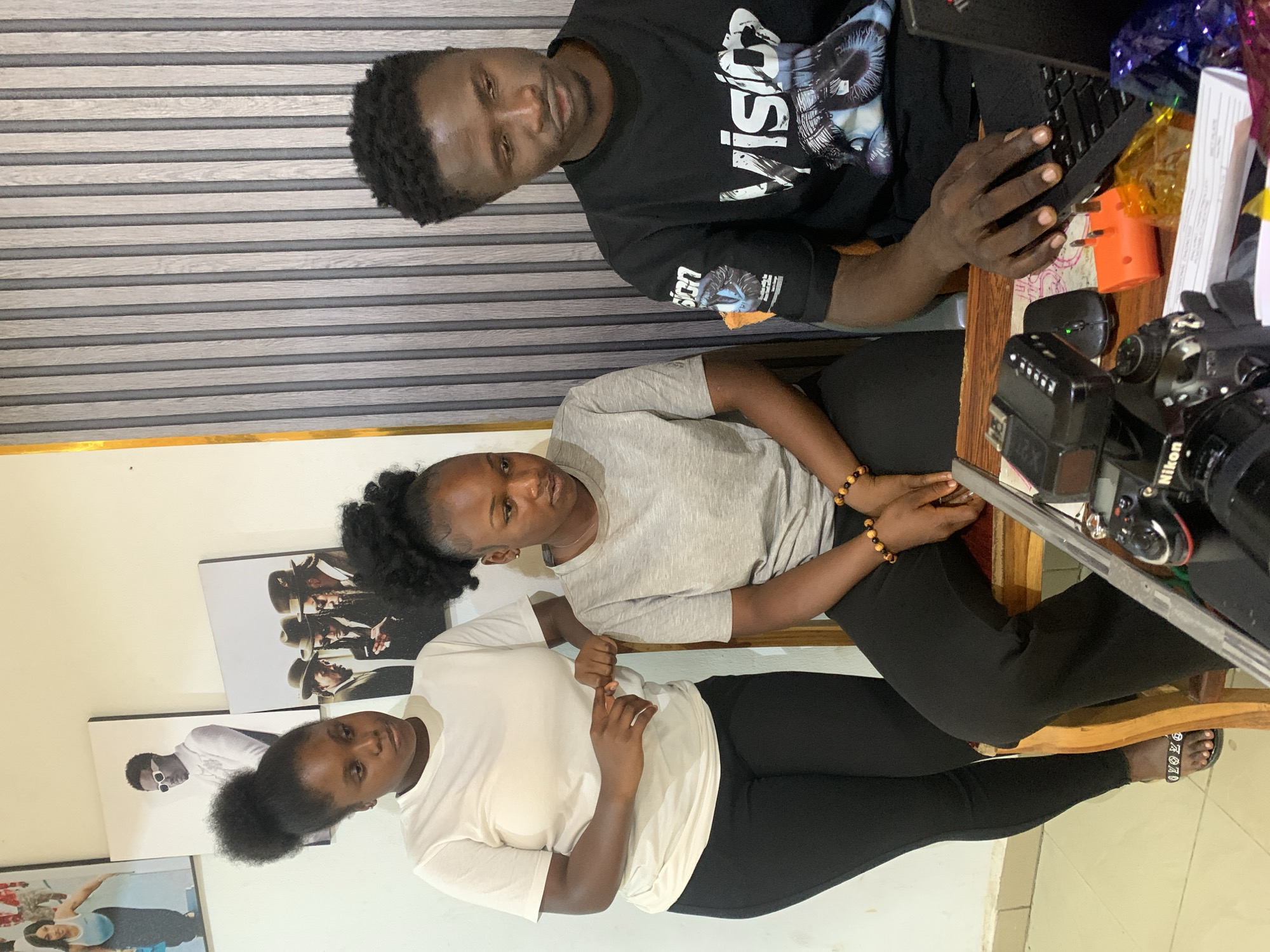 Three people are sitting at a wooden table in what appears to be an indoor office space. A man at the top of the frame is wearing a black t-shirt with "Vision" printed on it, and is using a laptop computer. Below him, a woman in a light gray t-shirt sits next to another woman in a white t-shirt. All three individuals appear to be looking at the camera. Black leggings are being worn by the woman in white and the woman in gray.
On the table, there is a camera with "Nikon" branding, a wireless computer mouse, and other assorted objects. The backdrop includes a wall with what appears to be horizontal gray and white panels at the top and some framed art prints on the wall.
Given the reference to Jos, Nigeria, it is possible that this scene is taking place in that city, but there's nothing in the image itself to verify the specific location.