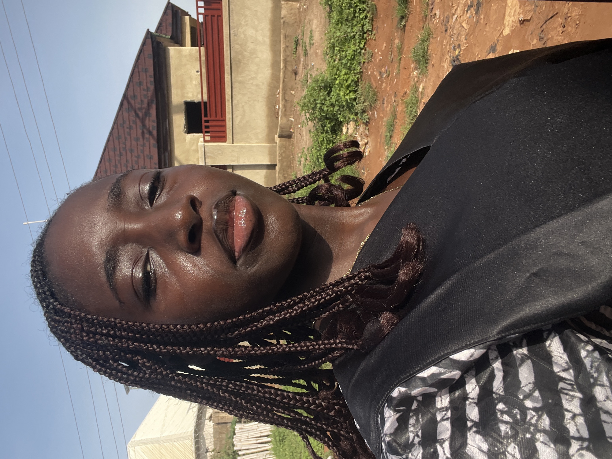 A person with dark skin and braided hair is photographed in a medium close-up shot. The person is wearing a black garment with a white and black patterned lining visible. The background includes a building with a reddish-brown roof and beige walls, patches of green vegetation, and brown soil. The sky appears light blue. The location is the city of Abuja, Nigeria.