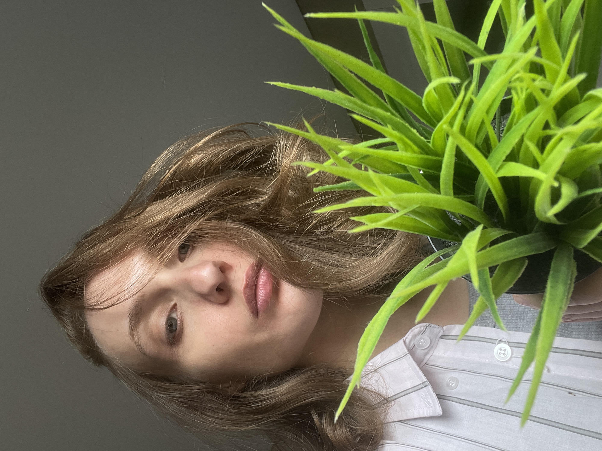 A woman with wavy, light brown hair is holding a small, artificial potted plant. She is wearing a light pink and white striped collared shirt. The background is a plain gray wall. The photograph is taken from a low angle, looking up at the subject. The image does not contain any information that would allow me to verify its location in Подшивалово, Russia.