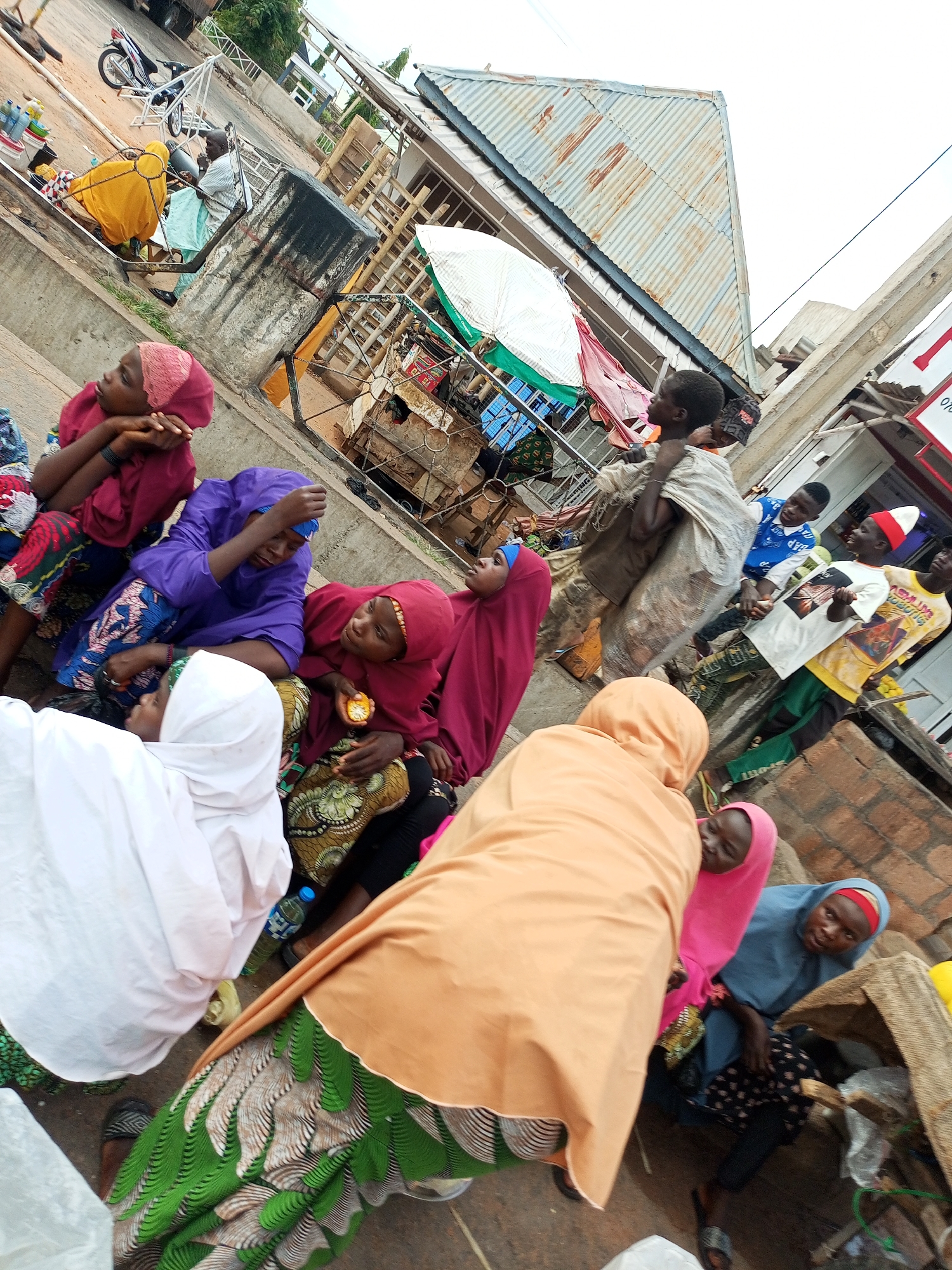 In Gombe, Nigeria, a group of people are gathered outdoors on a tiered stone structure. The overall scene is tilted, suggesting the photo was taken at an angle. In the foreground, several women and children are seated or kneeling. One woman in a white hijab is partially visible. Another woman wears a purple hijab and is looking down. A woman in a dark red hijab is holding an orange fruit. To her left, a woman in a bright orange hijab is facing away from the camera. A child wearing a pink hijab is visible next to the woman in orange. Further back, a person in a blue shirt is visible, and next to them, a child is being carried on someone's back. A man in a yellow t-shirt and a red and white hat stands near a building. A motorcycle is parked in the background to the left, and a blue plastic water bottle is on the ground near the seated individuals. The environment appears to be a bustling area with structures made of corrugated metal and concrete.