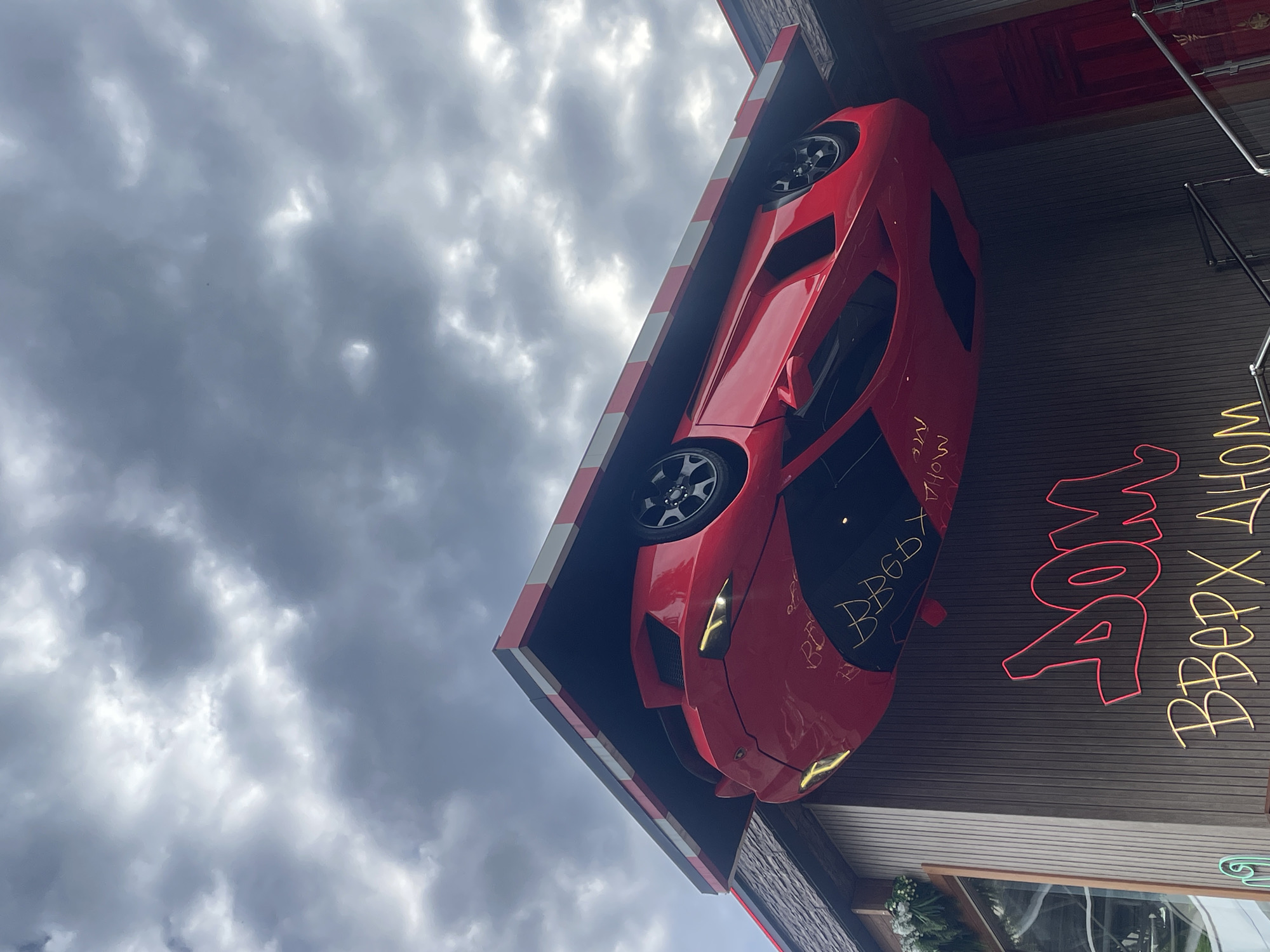 The image depicts an upside-down building in Sochi, Russia, featuring a bright red Lamborghini attached to its facade. The building is identified by a neon sign in Russian script that reads "ДОМ ВВЕРХ ДНОМ," translating to "House Upside Down." Graffiti in white marker is visible on the car's hood. The sky is overcast with gray and white clouds. The building's roofline has alternating red and white stripes. A small window with potted flowers is visible near the bottom edge of the image. The background includes additional architectural details and what appears to be a metal staircase attached to the building.