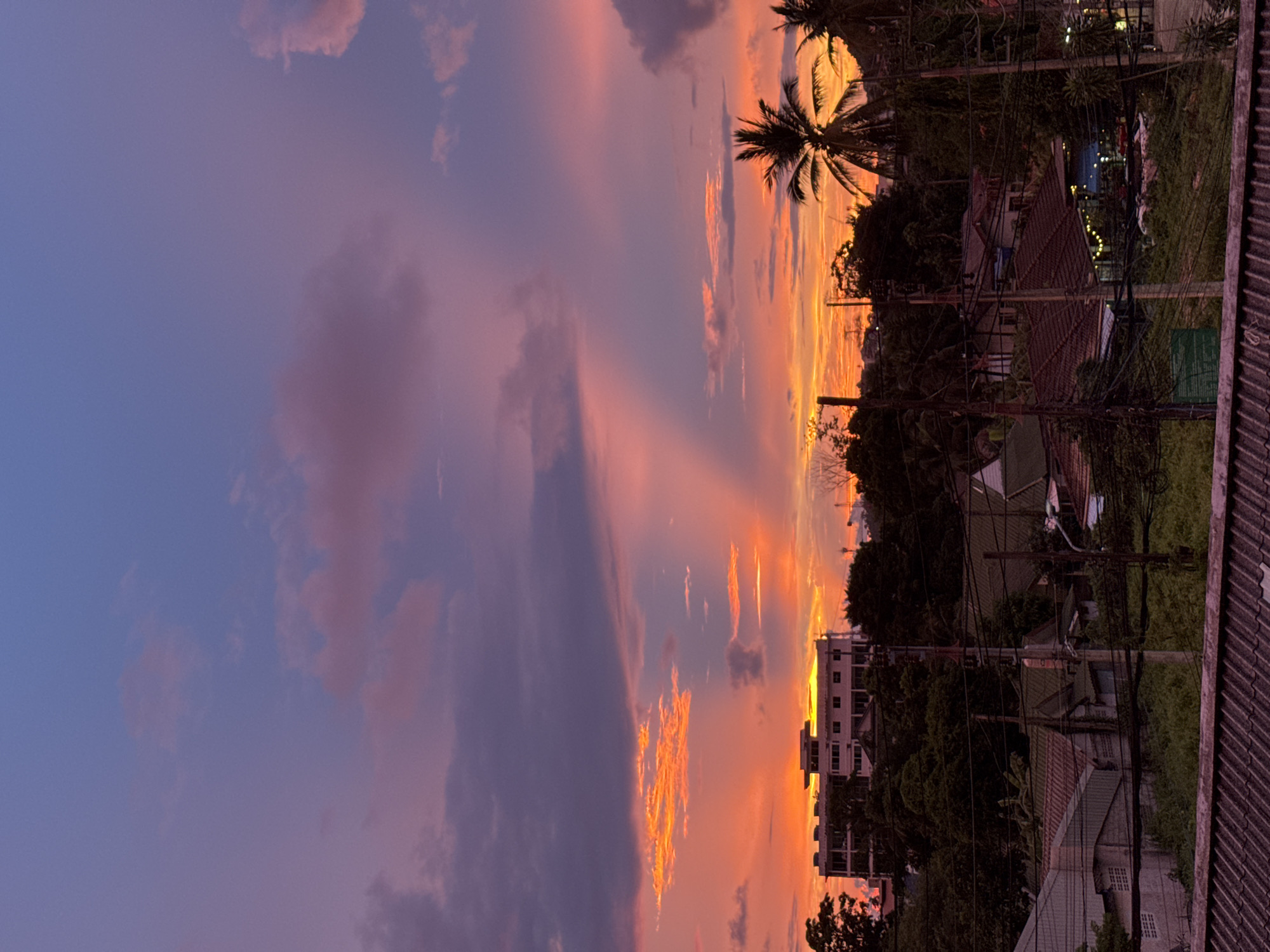 The image depicts a vibrant sunset over the city of Karon, Thailand. The sky is a dramatic canvas of deep blues, purples, and fiery oranges, with clouds catching the last rays of sunlight. In the mid-ground, a multi-story building with a flat roof and several windows stands amidst lush green trees. Below, a cluster of houses with terracotta roofs and various architectural styles are visible, some with lights on, suggesting evening activity. Palm trees, characteristic of tropical regions, punctuate the landscape. A network of electrical wires and poles crisscrosses the lower part of the scene, indicating the presence of infrastructure within the residential area. The foreground is dominated by a rooftop with visible texture. The overall scene captures a serene and picturesque tropical evening.