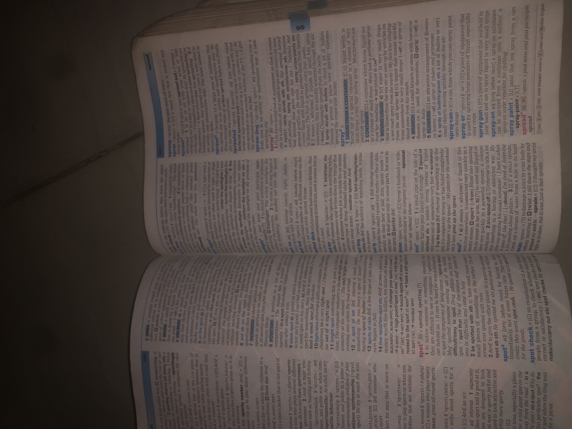 The image is a close-up, overhead shot of an open dictionary, focusing on pages detailing words starting with "sport" and "spraw." The dictionary pages are illuminated, showing crisp black text against a white background. The surrounding area is dark, suggesting the image was taken in a dimly lit environment. The dictionary itself appears to be a substantial volume, open to a page with a narrow, vertical blue marker on the right side. There are no people or any elements that would indicate a specific city or location like Jalingo, Nigeria. The content is solely text from the dictionary.