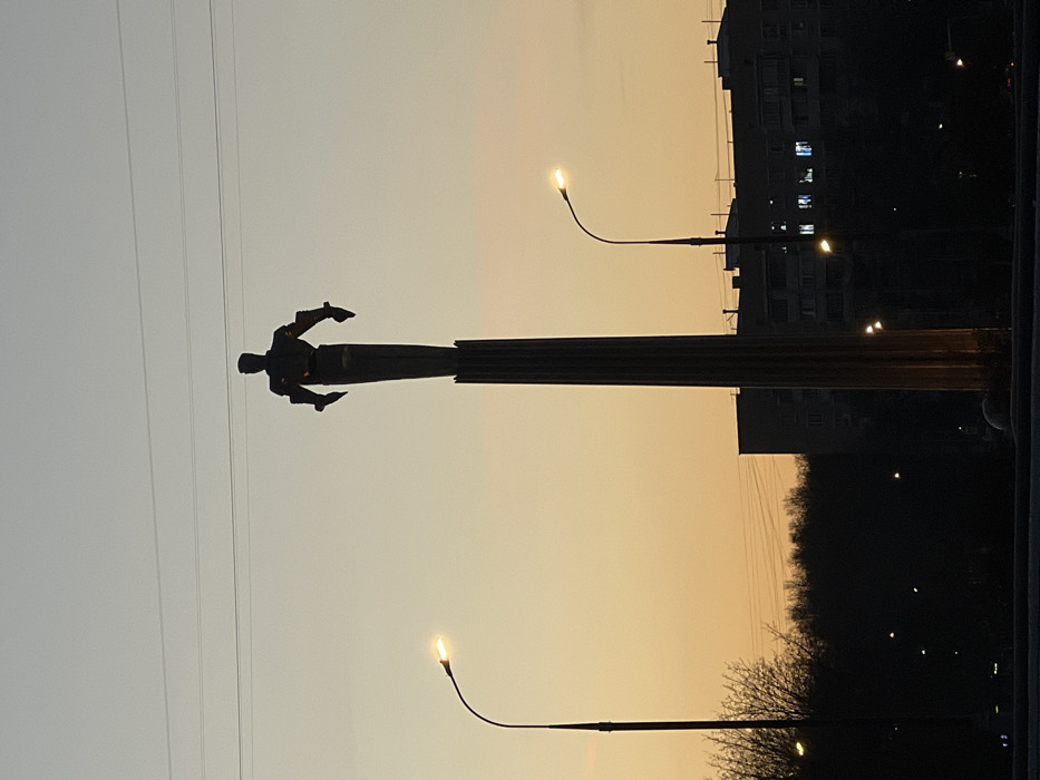 The image displays the Monument to the Conquerors of Space in Moscow, Russia, located near VDNKh on Prospekt Mira, during twilight. The central subject is the tall monument, silhouetted against the sky, featuring a prominent fluted column topped by a stylized dark figure representing a space explorer. The sky exhibits a gradient, transitioning from light orange-yellow at the horizon to pale grey-blue overhead. Two operational streetlights with bright, yellowish-white lamps are visible: one on the left and one on the right of the monument, both with curved standards. A dark, dense line of trees is present in the midground, primarily to the left of the monument. In the background, dark silhouettes of multi-story buildings are visible, with several windows emitting interior light. Multiple horizontal utility lines traverse the upper portion of the sky. No human figures or active vehicles are distinctly identified.
