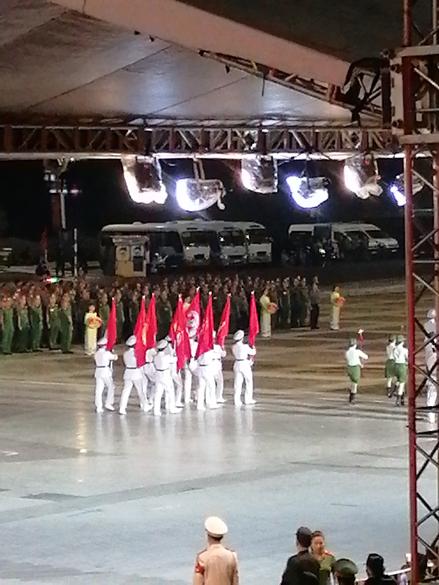 The image depicts a nighttime outdoor event, possibly a parade or ceremony, in Da Lat, Vietnam. The foreground shows a person in a light-colored uniform and cap facing away from the camera. Behind them, a group of individuals in dark uniforms are gathered. Further back, a formation of people in white uniforms and white caps are marching, many holding red flags. To the left, a larger crowd in green and dark uniforms is visible. Several white buses are parked in the background. The scene is illuminated by stage lights, casting a bright glow. The ground appears to be a wet, reflective surface, possibly from recent rain.