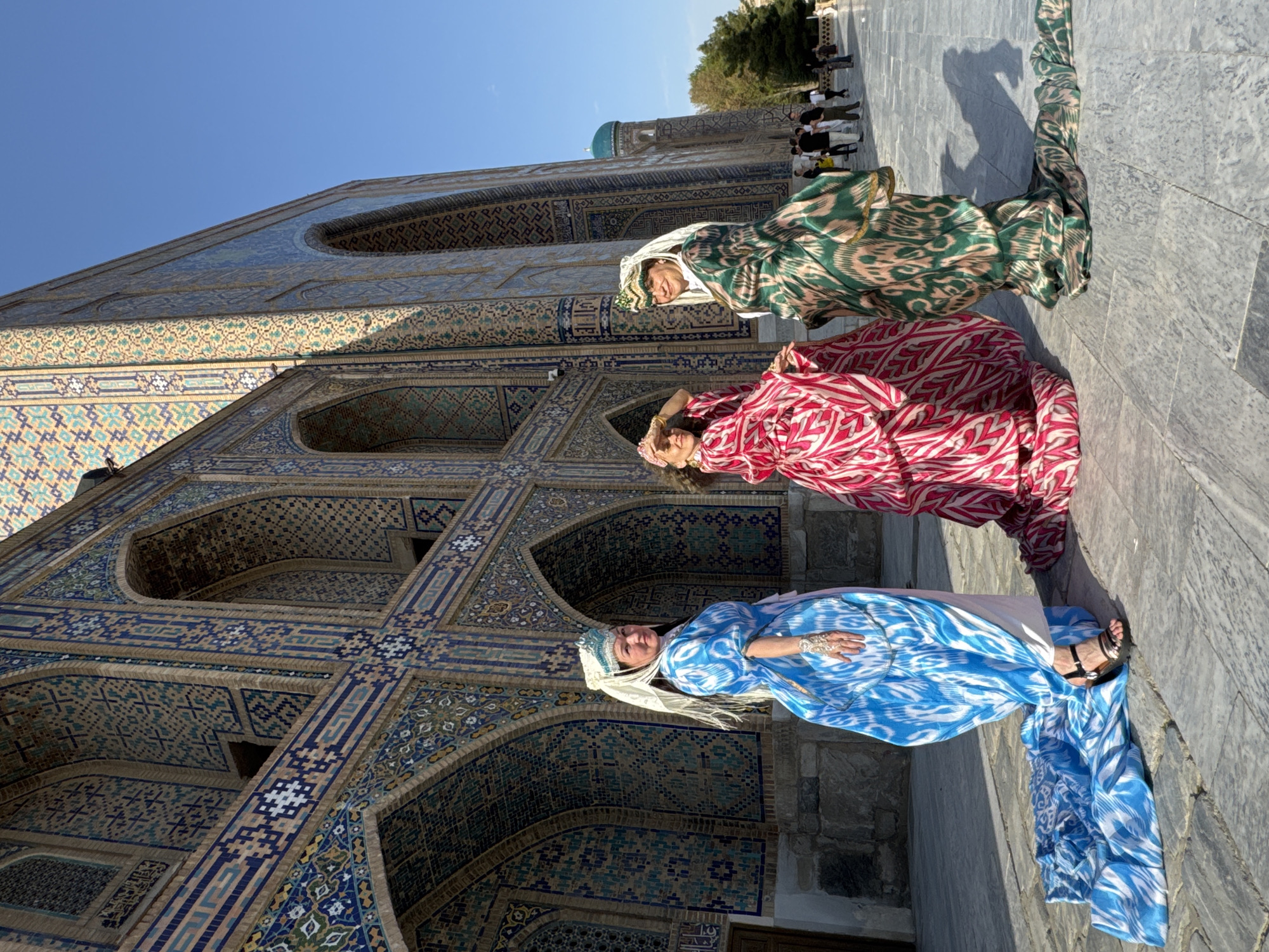 Three women stand in front of a building. The woman in the foreground is wearing a blue and white patterned dress and a white head covering. The woman in the middle is wearing a red and white patterned dress and head covering. The woman in the background is wearing a green and brown patterned dress and head covering. The building behind them is intricately tiled with blue, yellow, and white designs. The sky is blue. The women are standing on a stone walkway. People are visible in the background. The building is most likely the Registan in Samarkand City, Uzbekistan.