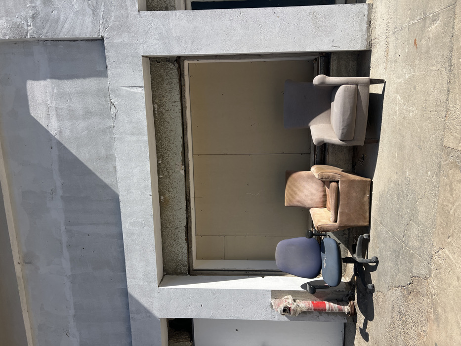 The scene depicts an outdoor urban environment in Tel-Aviv, Israel, under direct sunlight, indicated by sharp shadows. The background features a light grey, rough-textured building facade. A large, rectangular opening in the center of the facade is boarded up with light brown panels, secured within a recessed frame. To the left of this opening, a partially visible white door is set into a smaller recess. A red fire hydrant, partially covered with white wrapping material (possibly tape or plastic sheeting), is situated on the concrete ground adjacent to the white door.
Three chairs are arranged in a row on the cracked and discolored concrete ground in front of the boarded-up opening. From left to right:
1. A blue office chair with a high back, black armrests, and a black five-wheeled base. The seat and back show signs of wear and dust accumulation.
2. A brown or tan upholstered armchair with a textured fabric. This chair appears worn and somewhat soiled, positioned directly in front of the boarded opening.
3. A light grey or beige upholstered armchair, also showing signs of use, with visible wooden legs.
No individuals are present. No actions or interactions are depicted. The objects are stationary.