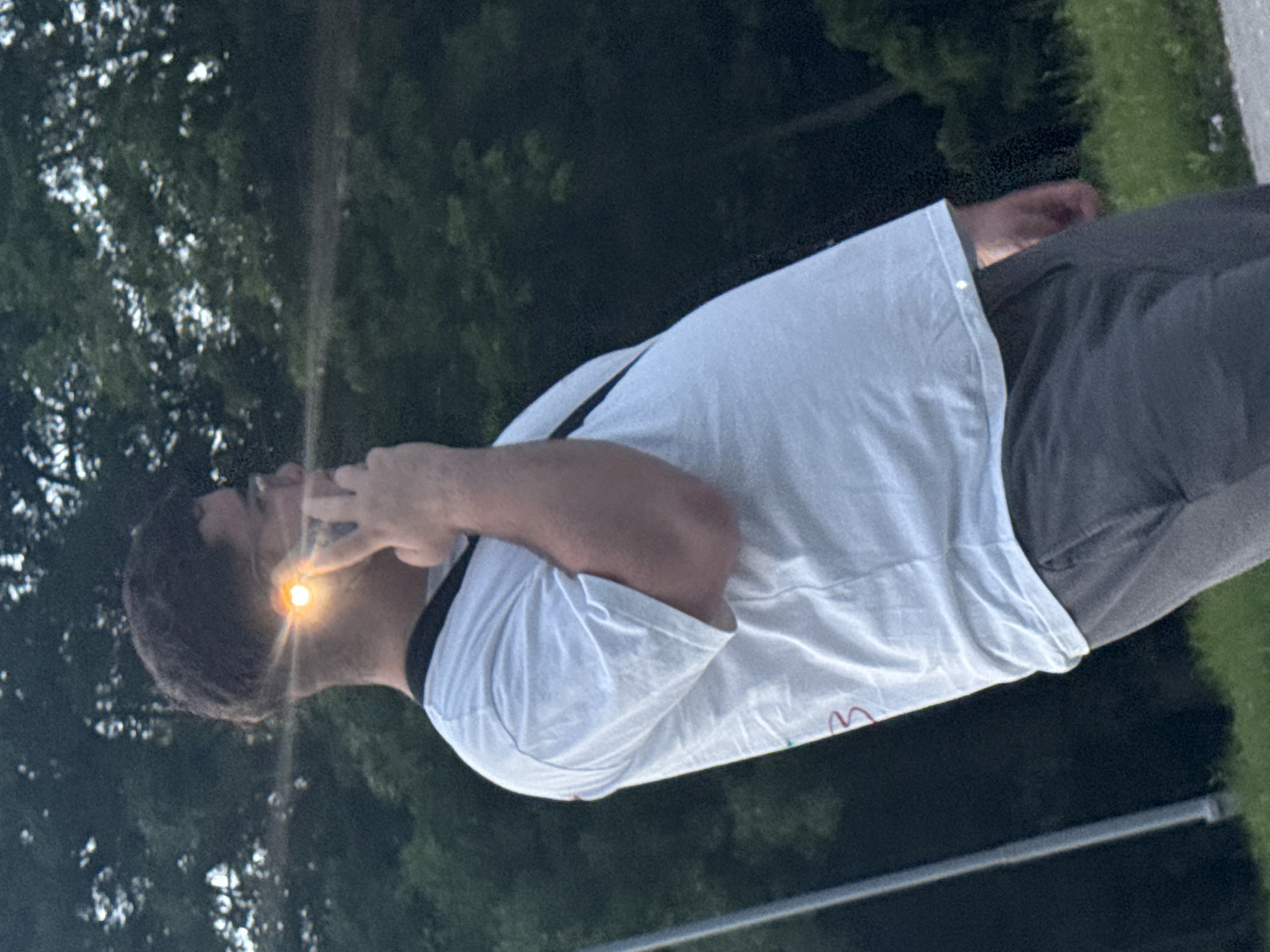 A man is standing outdoors, holding a mobile phone to his ear. The image seems to be taken at dusk or twilight, indicated by the dark sky and the bright light emanating from the phone screen. The environment appears to be a city park or similar green space. The precise location in the city of Pleven, Bulgaria, is not discernible based solely on this image.