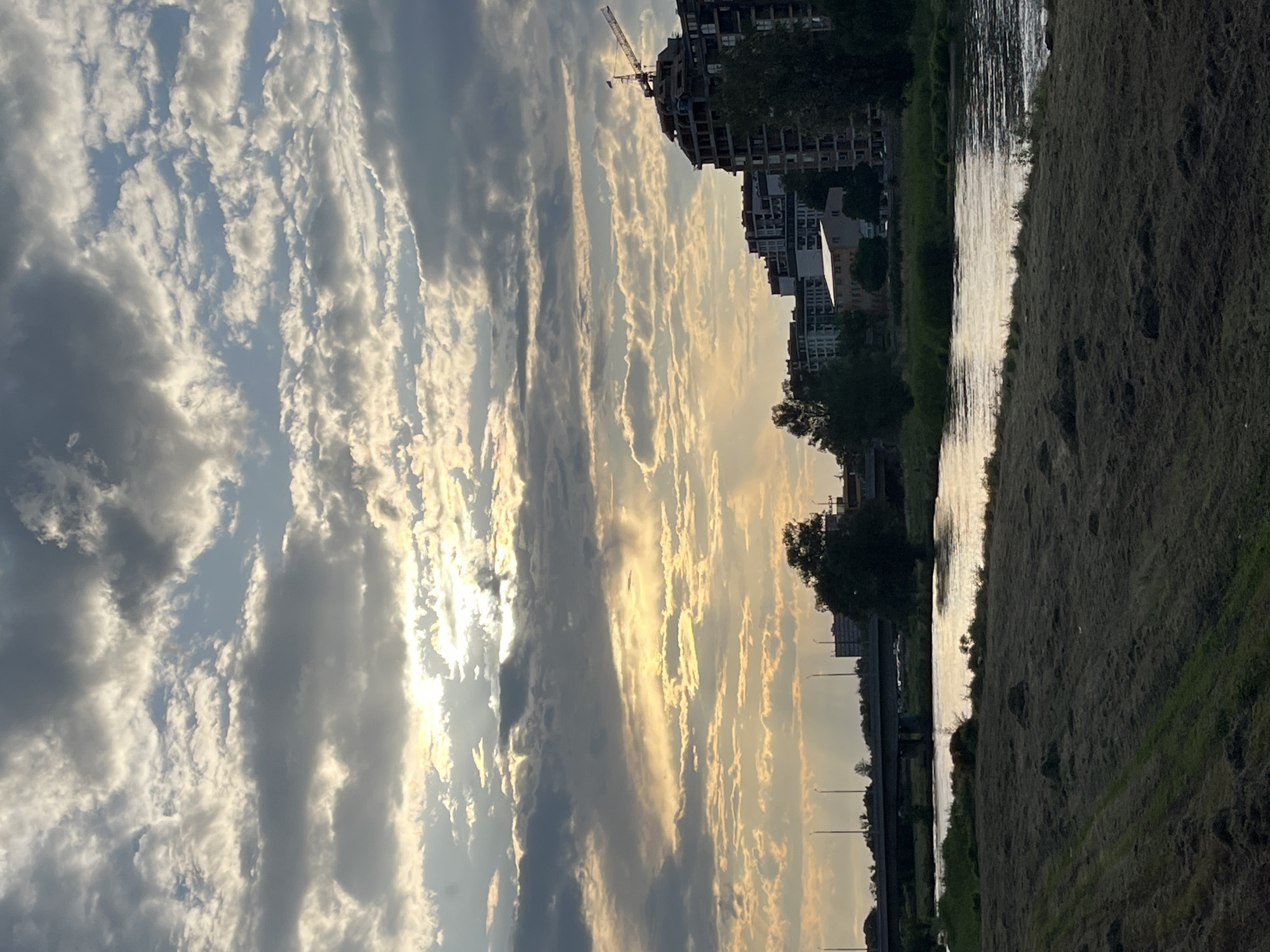 The image displays an expansive, partially cloudy sky over an urban riverside landscape, likely captured during sunrise or sunset in Plovdiv, Bulgaria. The sun, positioned towards the right of the frame, casts a golden, warm light that illuminates the edges of numerous elongated cloud formations, creating a striking visual contrast between bright, glowing segments and darker, grey cloud masses. Patches of clear, light blue sky are visible between the clouds.
A body of water, presumably a river, spans horizontally across the mid-ground, acting as a natural mirror for the sunlit sky. Its surface shimmers with reflections of the bright light, creating streaks of brilliance. On the immediate bank to the right of the river, there is an area of textured ground, covered with dry-looking grass and darker patches.
To the left of the river, an urban environment unfolds. Prominently in the upper left, a large building appears to be under construction, marked by a visible crane extending into the sky. Other buildings are seen further along the left bank, some partially obscured by trees that line the riverside. In the background, what appears to be a bridge or overpass structure is faintly visible, along with distant streetlights.
There are no people or animals visible, and no specific activities are taking place; the scene is a static depiction of the natural and built environment. The notable details include the dramatic lighting characteristic of dawn or dusk, the extensive cloud patterns, and the reflection of light on the water, all contributing to a tranquil and picturesque setting. No text is visible in the image.