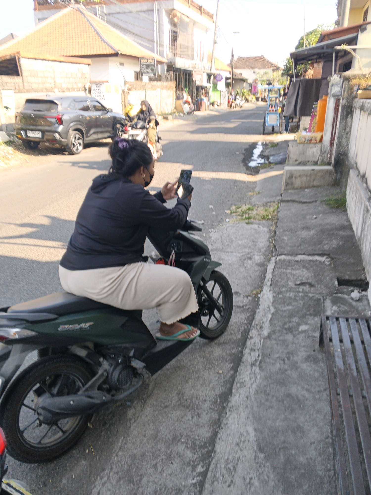 An over-the-shoulder, medium shot captures a woman on a dark green scooter from the perspective of someone behind and slightly to her left. The woman, wearing a black hoodie and light-colored pants, is looking down at her phone, which she holds in her right hand. Her left hand is on the scooter's handlebars. She is wearing green flip-flops. The scooter is parked on the right side of a narrow paved road in what appears to be a residential or commercial area of Sukawati, Indonesia. The road is lined with buildings, some with tiled roofs, and a sidewalk on the right. Sunlight casts shadows on the road, indicating it's daytime. In the background, a gray SUV is visible on the left side of the road, and another person on a scooter is further up the street. A blue and white cart is also present on the right side of the road.