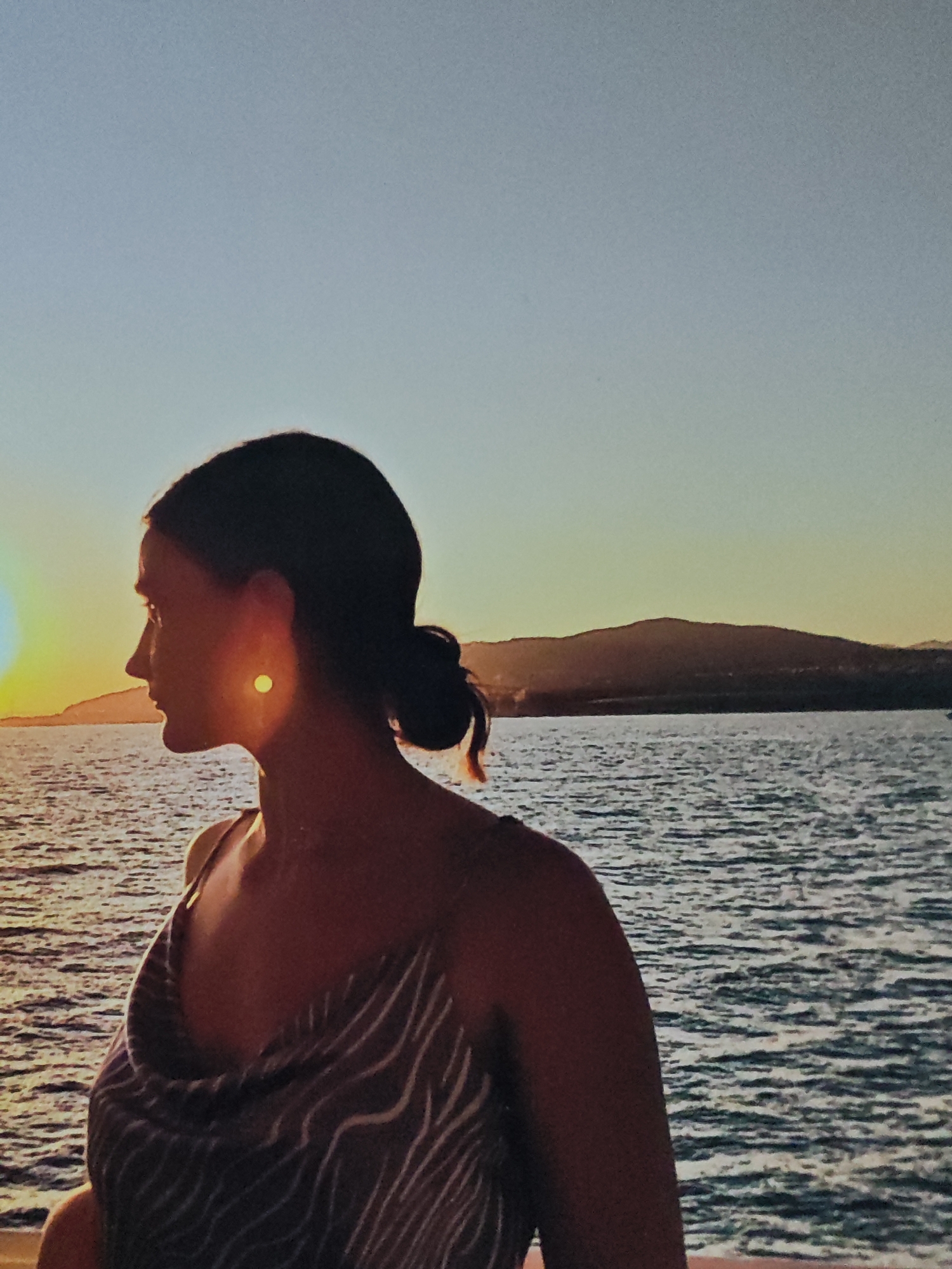 The image depicts a profile of a woman with dark hair pulled back in a bun, looking out at a body of water during sunset. The sun is visible on the left side of the frame, casting a warm glow and creating reflections on the water. In the background, hills are silhouetted against the sky. The woman is wearing a patterned top. The scene suggests an outdoor setting, possibly a coastal area, with calm water and a clear sky. The location is identified as Unknown, Unknown.