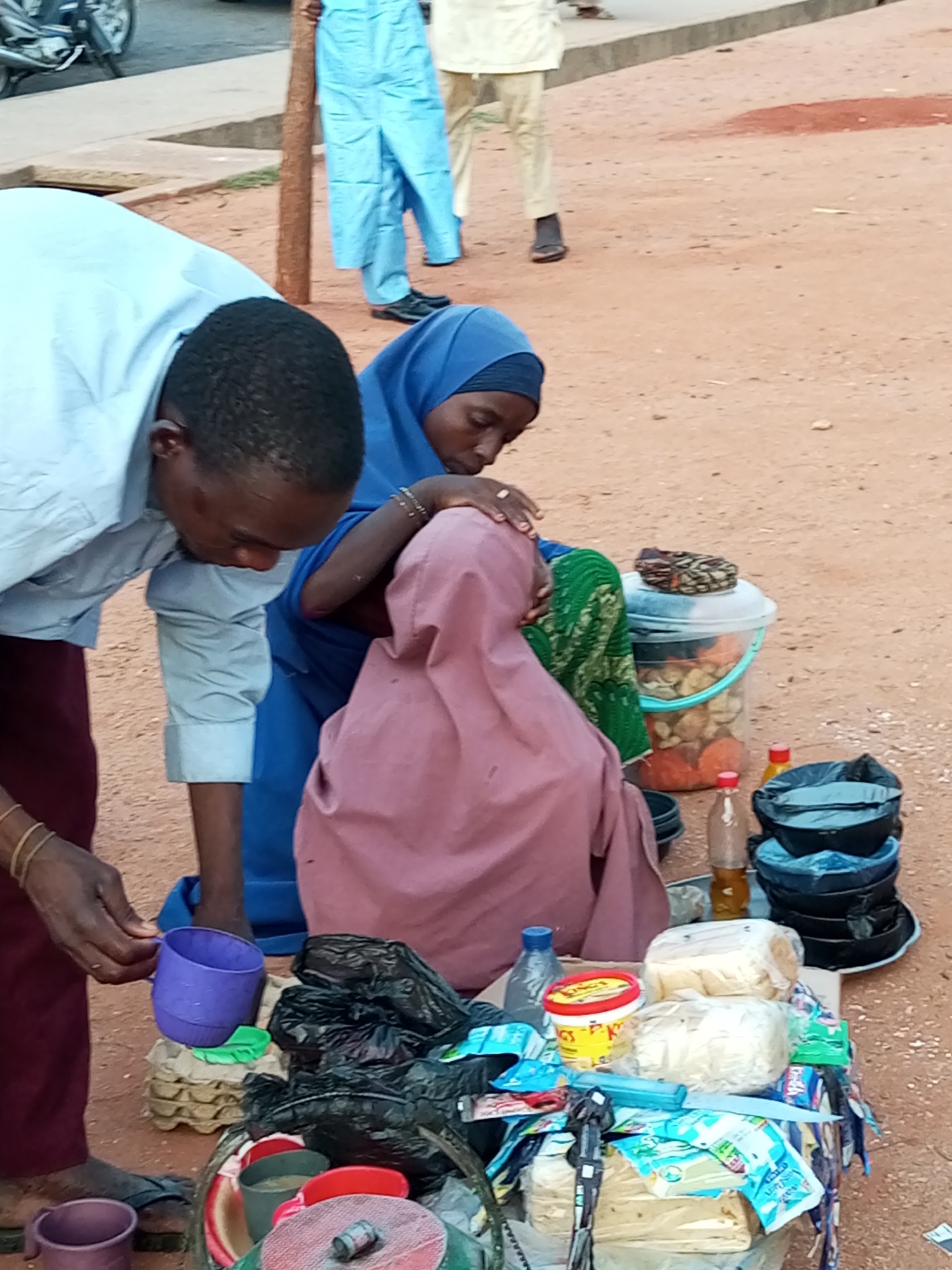This image depicts a street scene in Gombe, Nigeria, likely a marketplace or roadside vending area. In the foreground, a man in a light blue shirt and burgundy pants is bending over, holding a purple cup. To his right, a woman wearing a blue hijab and a green patterned garment is seated, also bending over. Another person, covered in a pink veil, is seated between them.
On a table in front of the seated individuals, various items are displayed for sale or use. These include packaged food items, possibly bread or snacks, wrapped in plastic. There are also several plastic containers, including a clear bucket filled with what appears to be food items like nuts or dried fruit, and stacks of black and blue plastic bowls. Bottles of liquid, one with a yellow contents, are also present. Bags, some black and some possibly containing produce, are scattered around the table.
In the background, two individuals are standing. One is wearing light blue traditional attire, and the other is in cream-colored clothing and light brown pants. A tree trunk stands to the left of the standing figures. The ground is composed of reddish-brown dirt, typical of the region. A motorcycle is partially visible on the far left, suggesting it is parked nearby. The overall impression is a candid snapshot of daily commerce and life in Gombe.