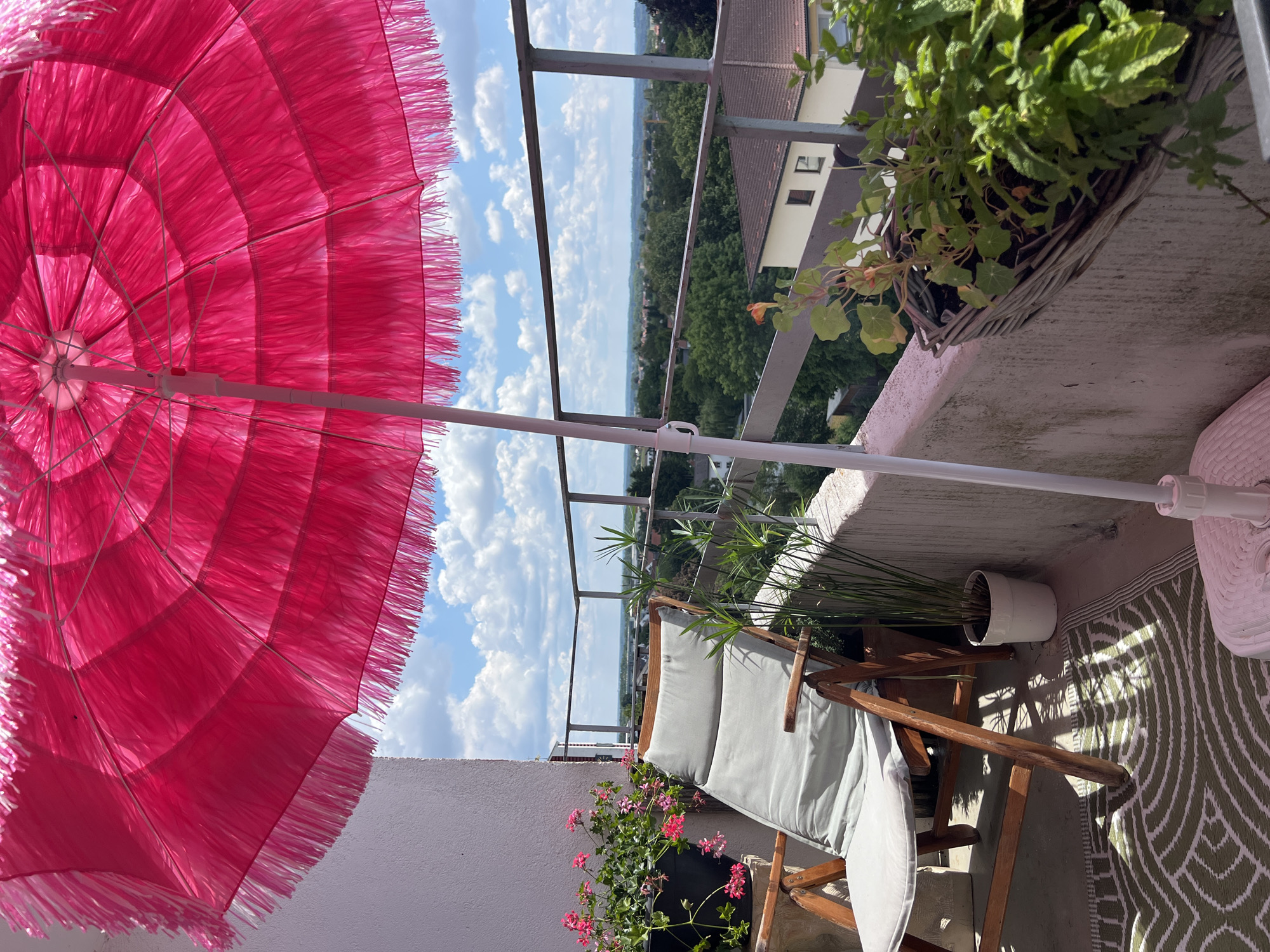 The image depicts a balcony in Mühldorf am Inn, Germany, on a sunny day with scattered clouds. In the foreground, a vibrant pink umbrella with fringe hangs overhead, casting shadows on the scene. To the left, a wooden lounge chair with a light gray cushion is positioned on a patterned rug. Beside the chair, a potted plant with green, reed-like leaves stands in a white pot. Further to the left, a black planter overflows with pink flowers.
A railing divides the balcony from the view beyond, which encompasses a lush green landscape of trees and houses, suggesting a residential area within the city. In the middle ground, a wicker planter holds various green plants, including what appears to be nasturtiums with orange flowers. The overall impression is one of a peaceful, outdoor seating area prepared for relaxation.