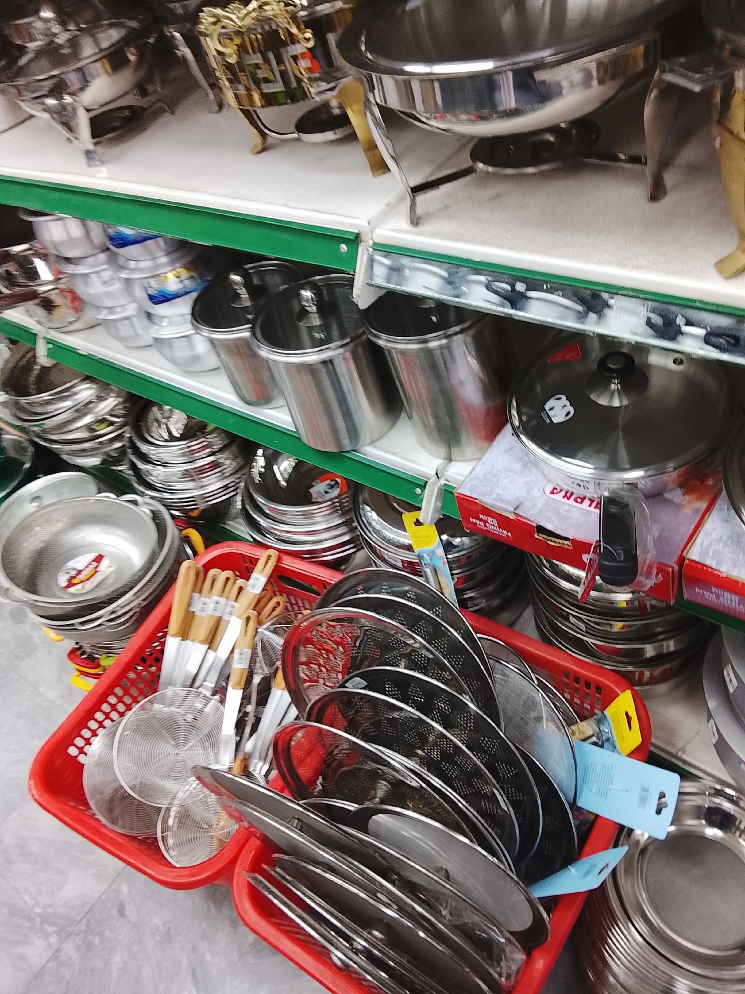 The image depicts a retail display in Gulgasht Colony, Pakistan, showcasing kitchenware. The scene is a close-up shot, featuring shelves stacked with various stainless steel items. On the top shelf, there are chafing dishes and warming pans. The middle shelf contains pots and pans, some with lids. In the foreground, a red plastic basket is overflowing with strainers and slotted spoons, also made of stainless steel. The floor appears to be tiled. There are no people visible in the frame. The objects suggest a store specializing in kitchen supplies or a general store with a kitchenware section.