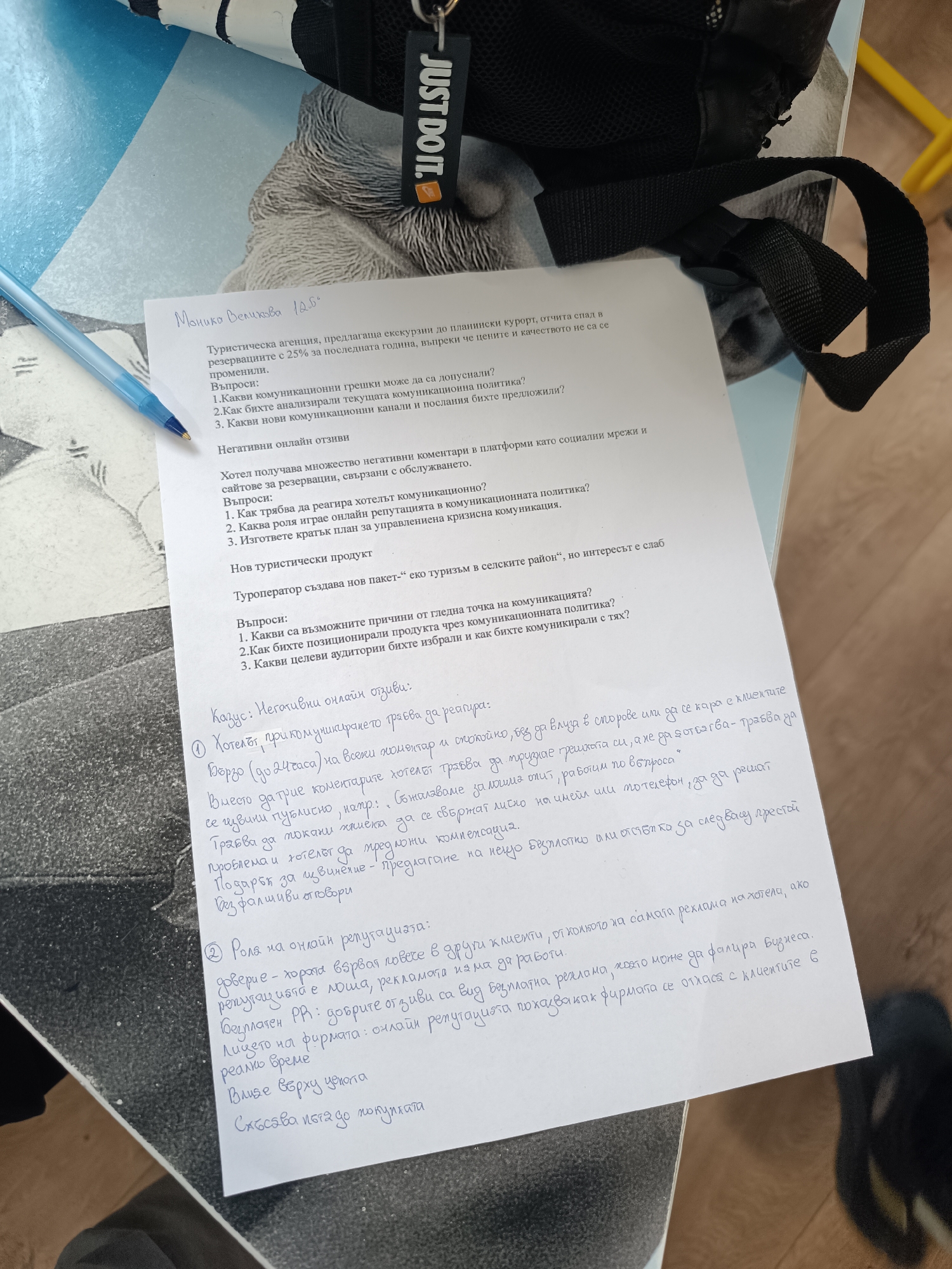 A top-down shot reveals a handwritten document titled "Tourism Agency" in Bulgarian, placed on a surface with a blue and white pattern. The document appears to be a case study or assignment, containing questions about communication strategies, negative online reviews, and a new tourism product related to eco-tourism. A blue ballpoint pen rests near the top of the document. A black backpack strap with the "JUST DO IT." logo is visible in the upper-right corner of the frame. The location, Pleven, Bulgaria, is not discernible from the visual information provided.