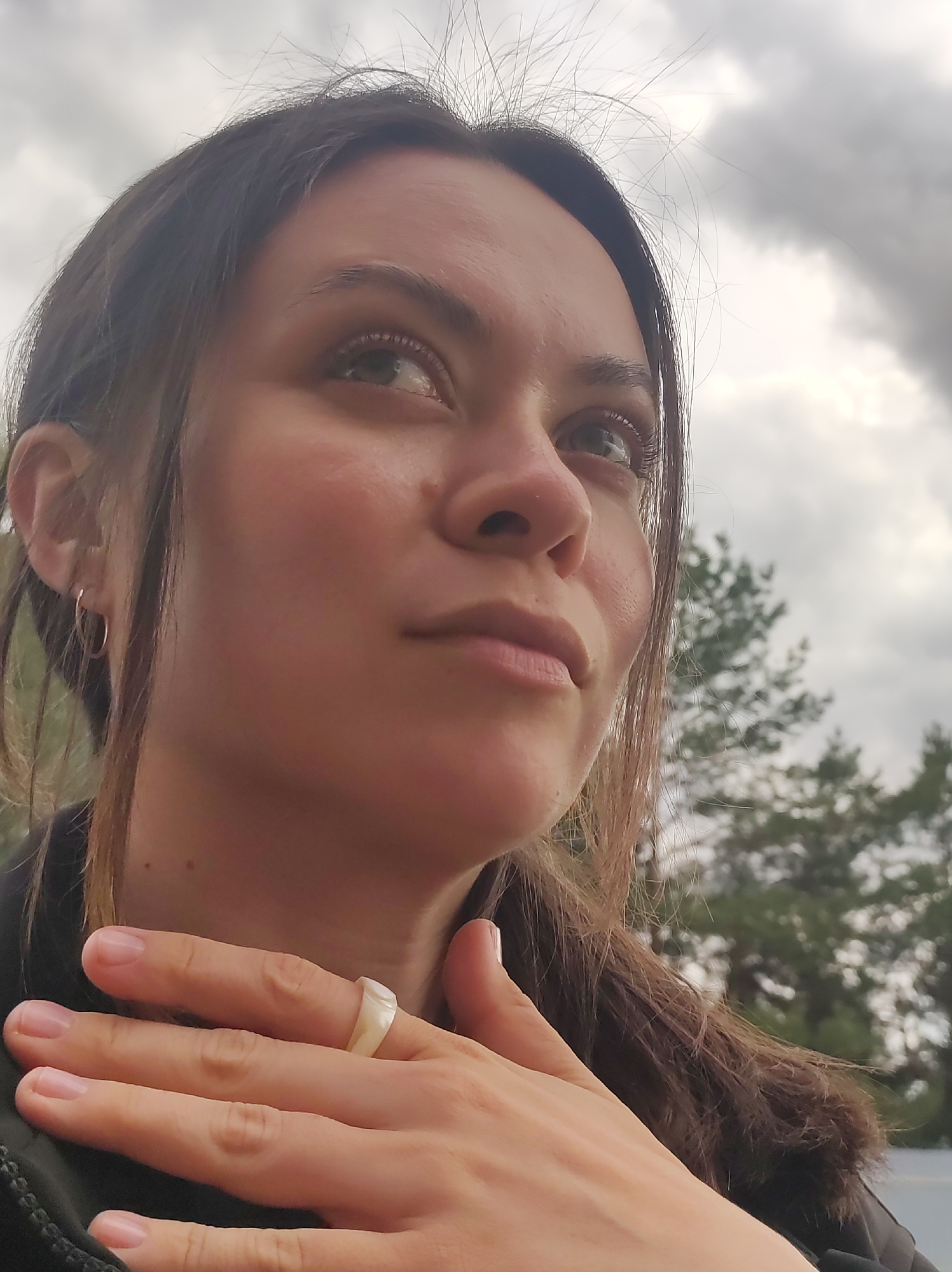 This is a close-up, eye-level portrait of a young woman taken outdoors in Фоки, Russia, on an overcast day. She is captured from the chest up, looking upwards and slightly to her right with a thoughtful or contemplative expression. Her dark brown hair is pulled back, with fine, wispy strands framing her face. She has light-colored eyes, natural complexion, and a few small moles visible on her cheek and neck. A small, simple gold-toned hoop earring adorns her left ear.
Her right hand is gently placed on her chest or collarbone area, and she is wearing a distinct, chunky, light-colored ring – possibly white or translucent plastic or stone – on her right ring finger. She is dressed in dark clothing, likely a jacket, with a hint of dark, possibly fur-lined, material visible near her right shoulder.
The background is softly blurred, dominated by a cloudy, gray sky. Below the clouds, dark green coniferous trees are visible, suggesting an outdoor, natural or wooded setting. The lighting is soft and diffused due to the overcast conditions. The overall mood conveyed by the image is calm and introspective. There is no visible text in the image.
