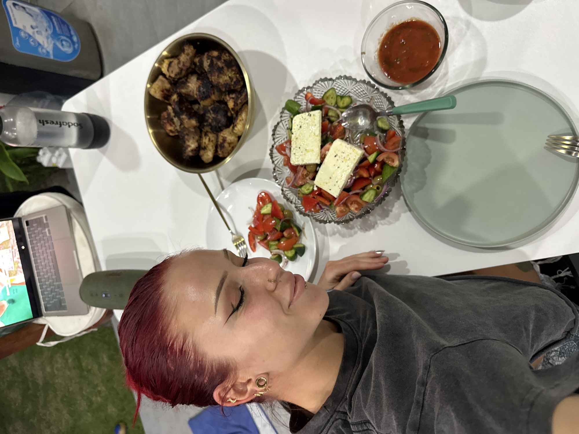 A person with red hair, multiple ear piercings, and a nose ring is seated at a white table. The table holds a laptop computer, a green portable speaker, a bottle of soda, a container, and food items including a bowl of grilled meat, a salad with feta cheese, a small plate of salad, and a bowl of sauce. A green plate with a fork is also visible. The person is wearing a gray shirt. The setting is North Kuta, Indonesia.