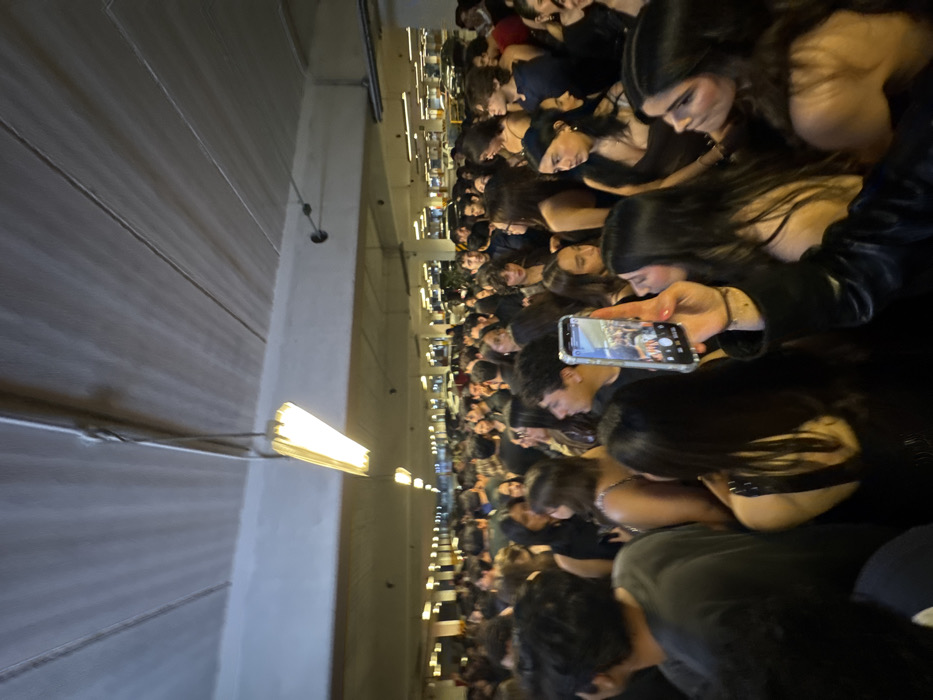 A dense crowd of people is packed into a large hall, likely an auditorium or performance venue. Most appear to be young adults, primarily with dark hair. Many are looking downward, possibly at a stage or performance out of view. A few individuals are holding up their phones, seemingly recording or taking pictures of the event. The lighting in the hall is predominantly provided by long, fluorescent lights hanging from the ceiling. The overall atmosphere suggests a lively but packed indoor event.