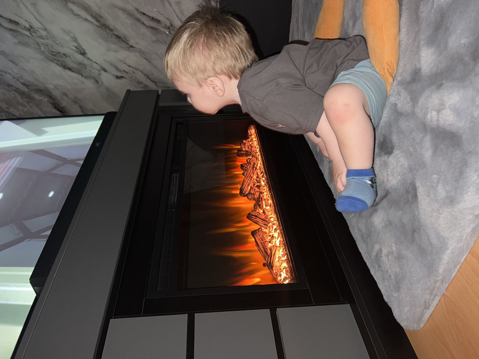 A toddler in Perm, Russia, wearing a gray shirt, light blue shorts, and a blue sock, leans forward to look into an electric fireplace. The fireplace has a black frame and artificial logs illuminated with orange light. A plush blanket in gray and orange rests on a surface near the child. A television is visible on the left side of the image, displaying an interior scene. A wall with marble-like patterns is in the background.