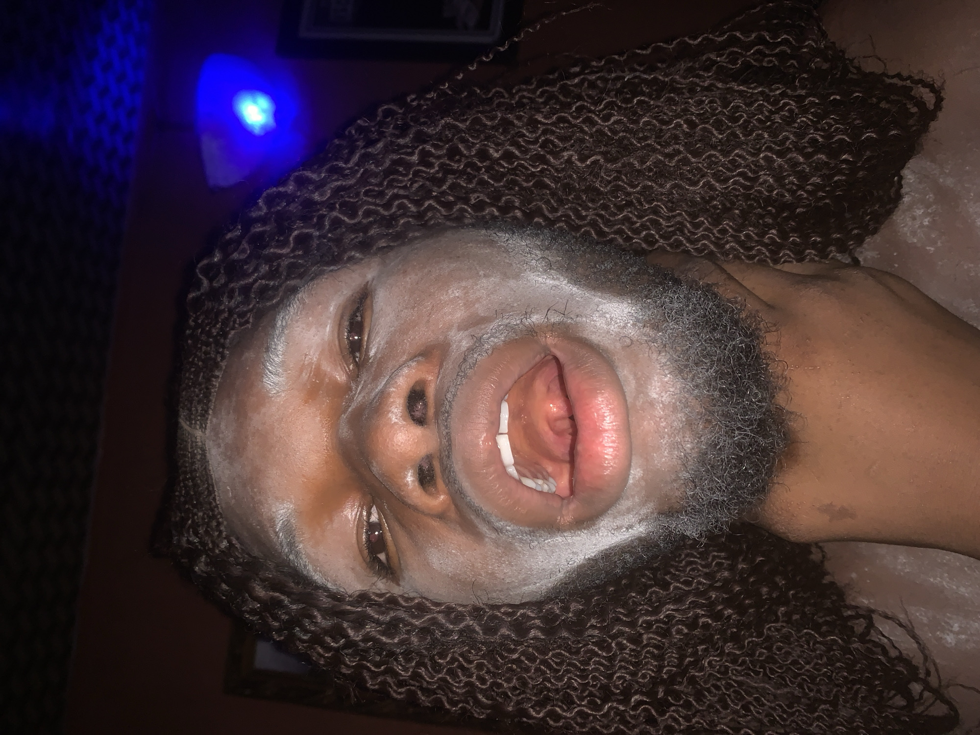A close-up portrait features a person with dark skin, oriented sideways in the frame. The individual has long, dark, tightly curled or wavy hair, possibly styled as a wig or extensions, which frames their face and extends around their head. Their face, including the forehead, cheeks, and chin, is coated with a light-colored substance, resembling a face mask or powder. A beard and mustache are also visible, partially covered by this application.
The person's mouth is wide open, revealing their white teeth and the interior of their mouth. Their eyes are open and gaze directly towards the viewer. In the upper left portion of the background, a distinct bright blue light source emits a glow, illuminating a small area. The overall scene appears to be dimly lit, suggesting an indoor setting during the evening or night. The background is dark and largely indistinct, with subtle textural elements. The expression, with the wide-open mouth and direct eye contact, conveys a sense of engagement or playfulness. No visible text is present in the media. The image's context is Onitsha, Nigeria.