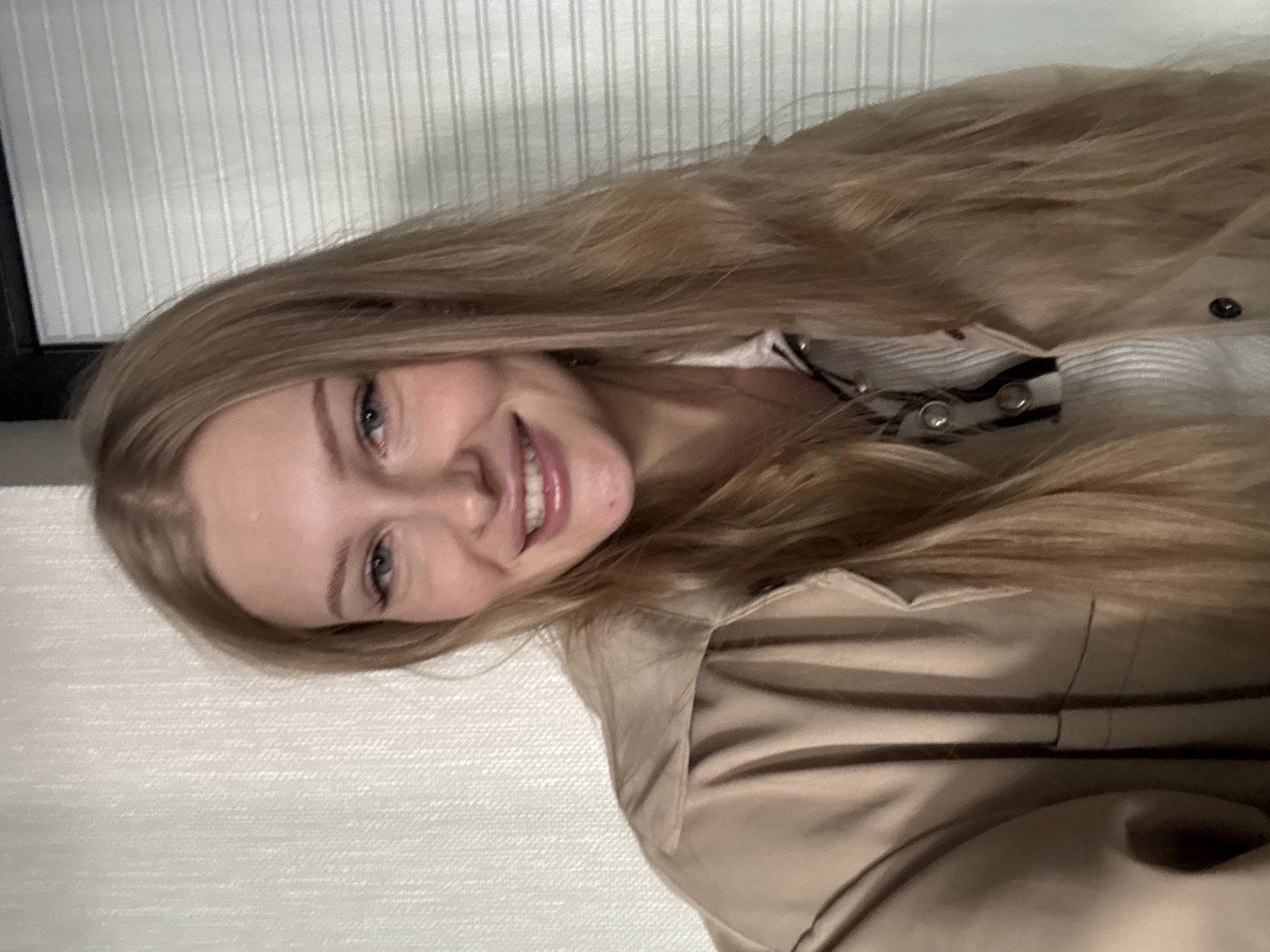 A young woman with long, blonde hair is captured in a close-up, head-and-shoulders shot, indoors. She is looking directly at the camera with a slight smile. Her eyes are blue, and she has fair skin with a few freckles. She is wearing a light-colored trench coat over a white ribbed shirt with dark stripes and silver buttons. The background features a textured, light-colored wall on the left and a horizontally slatted, light-colored surface on the right. The lighting is soft and appears to be coming from the front. There is no visible indication of the city of Perm, Russia, in the image.