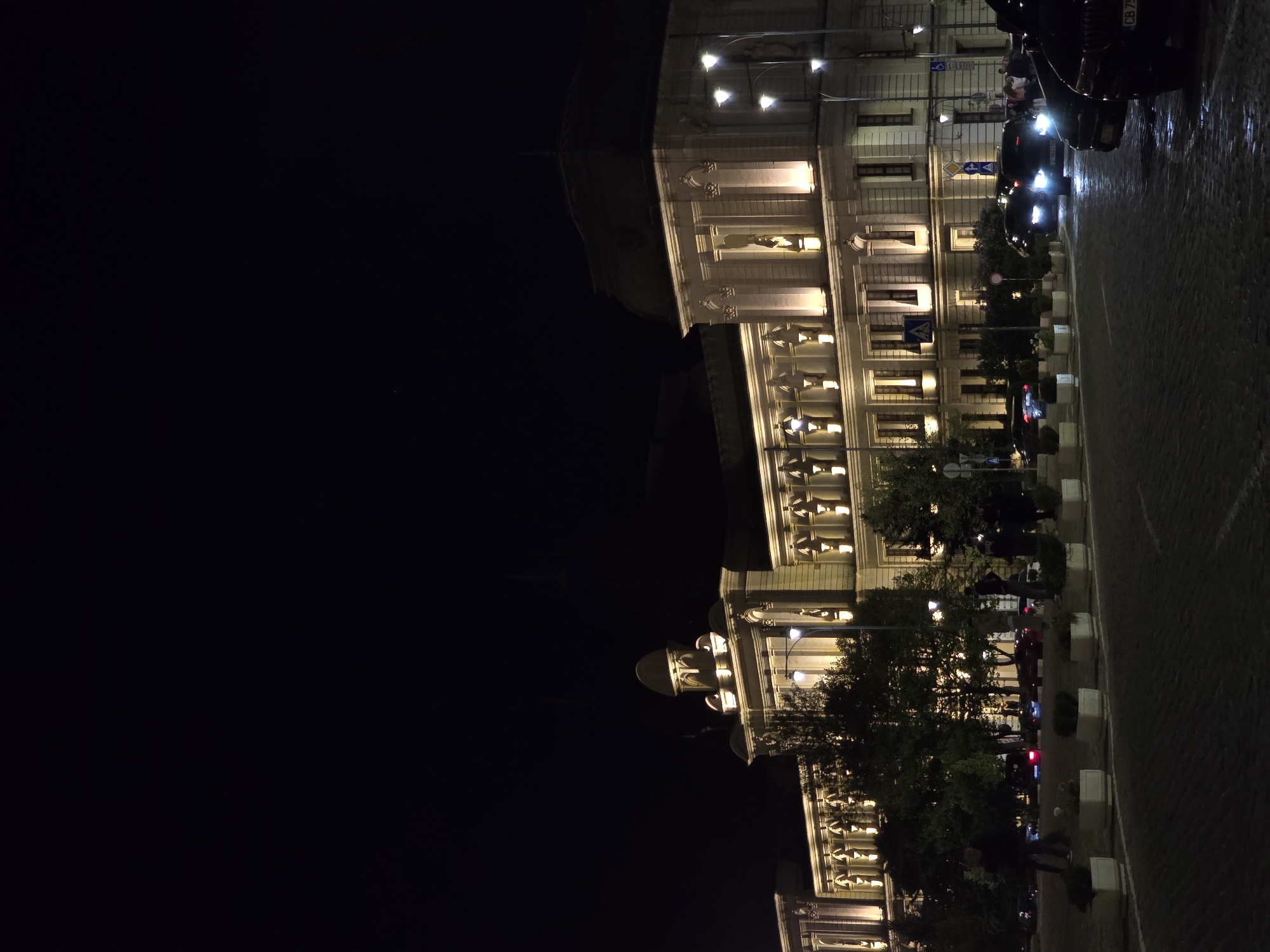 At night, a tall building with illuminated windows stands beside a cobblestone street in Sofia, Bulgaria. Cars drive on the street, and people walk nearby. The sky is dark.