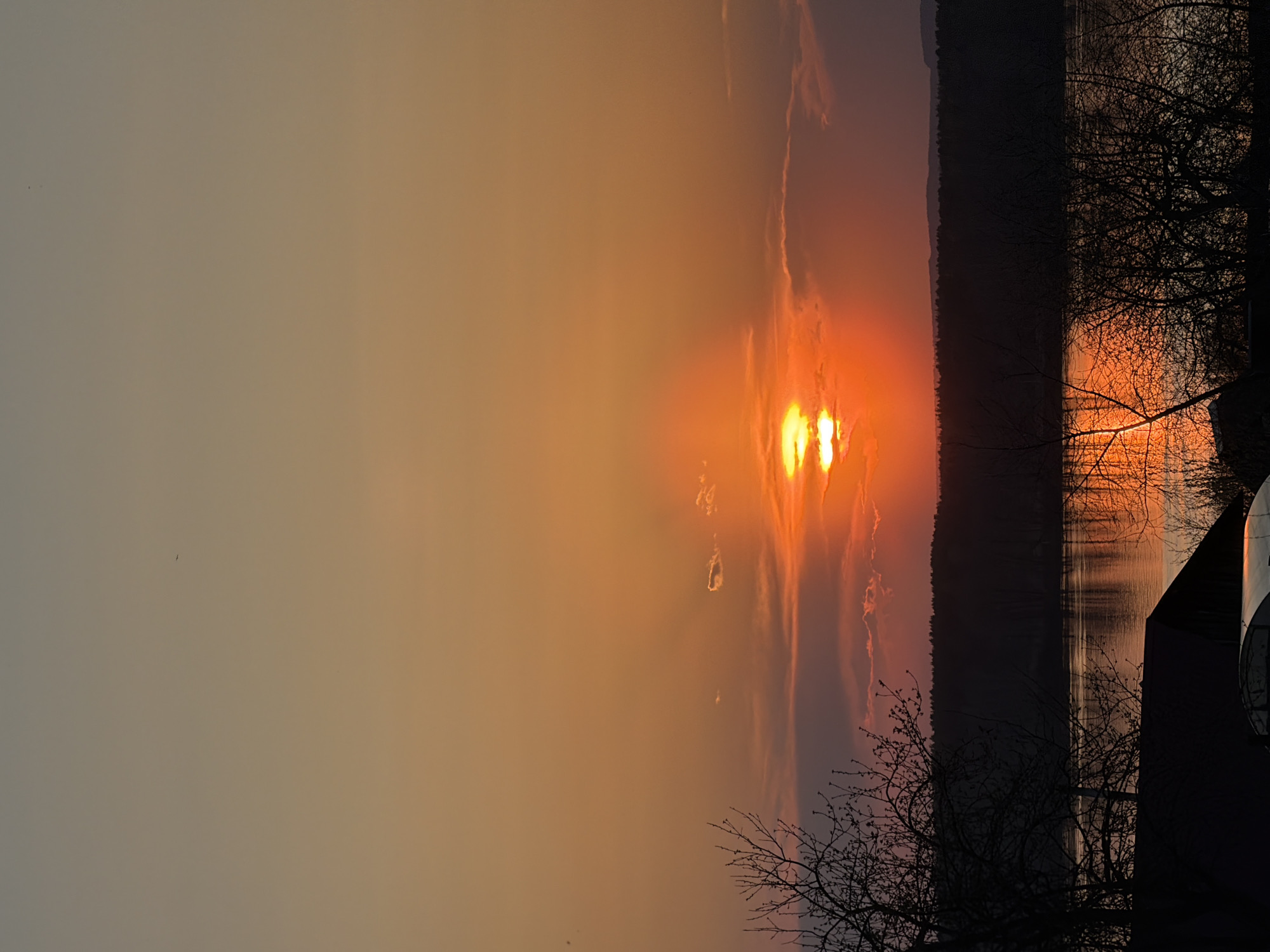 The image captures a sunset over a body of water, likely near Chaikovsky, Russia, evidenced by similar landscape features. The sun is setting near the horizon, casting an orange glow across the sky, with some clouds visible. In the foreground, silhouetted bare trees frame the view, and a dark structure, possibly a building, is partially visible on the right side of the image. The water reflects the colors of the sunset, creating a mirror image effect. A dark strip of land or trees runs horizontally across the middle of the image, separating the sky from the water.