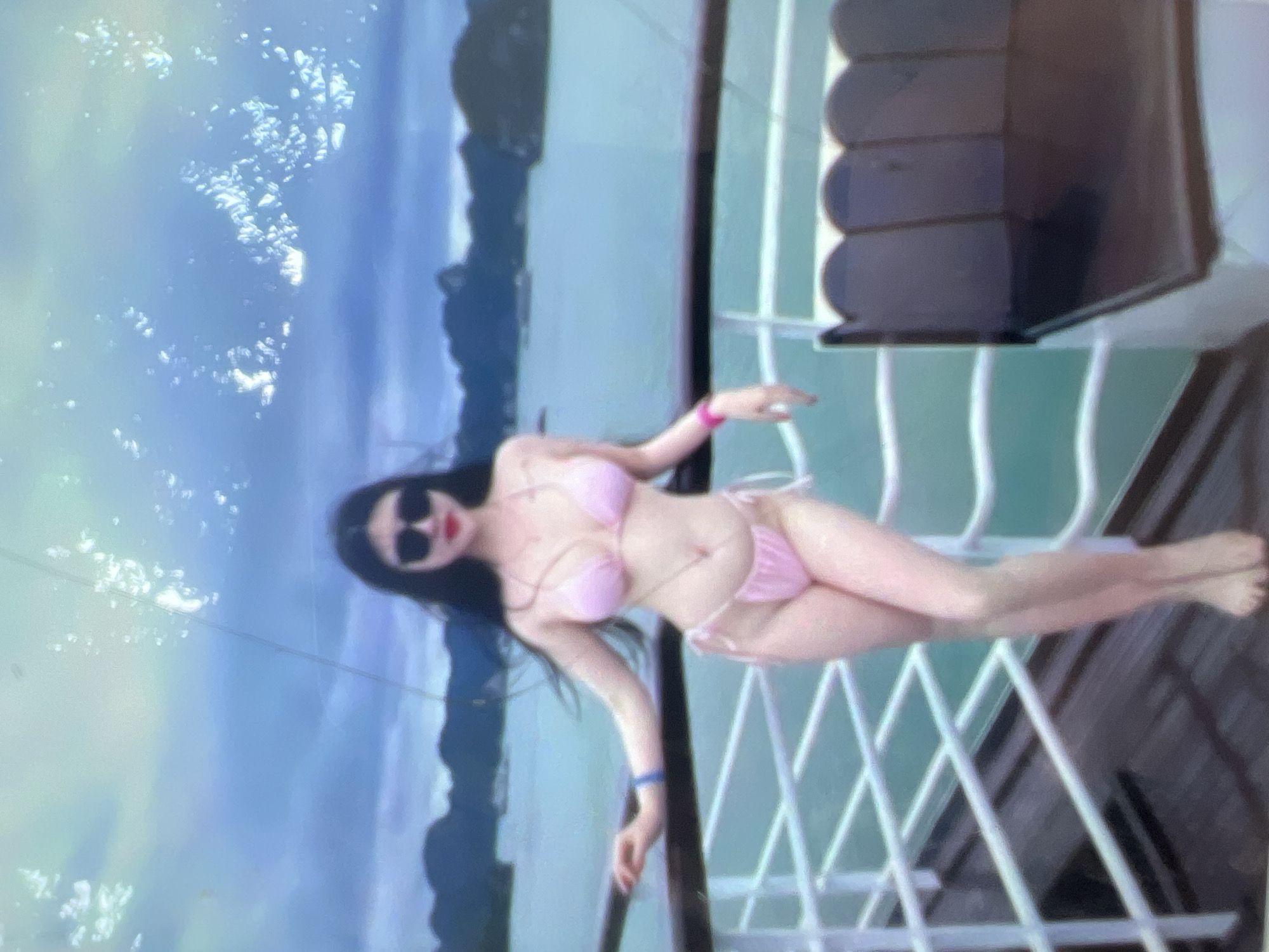 A full-body, waist-up shot features a woman in a pink bikini standing on the deck of a boat. She has long, dark hair and wears sunglasses and red lipstick. Her left arm is extended, resting on a railing, while her right arm is bent at the elbow. The boat deck has white railings and dark wooden flooring. In the background, a turquoise body of water is visible, with green, mountainous islands dotting the horizon under a cloudy, pale blue sky. The image is somewhat blurry and has a grainy texture with bright, white flecks scattered across the top half. There is no indication of Kara-Dıykan, Kyrgyzstan in the image.