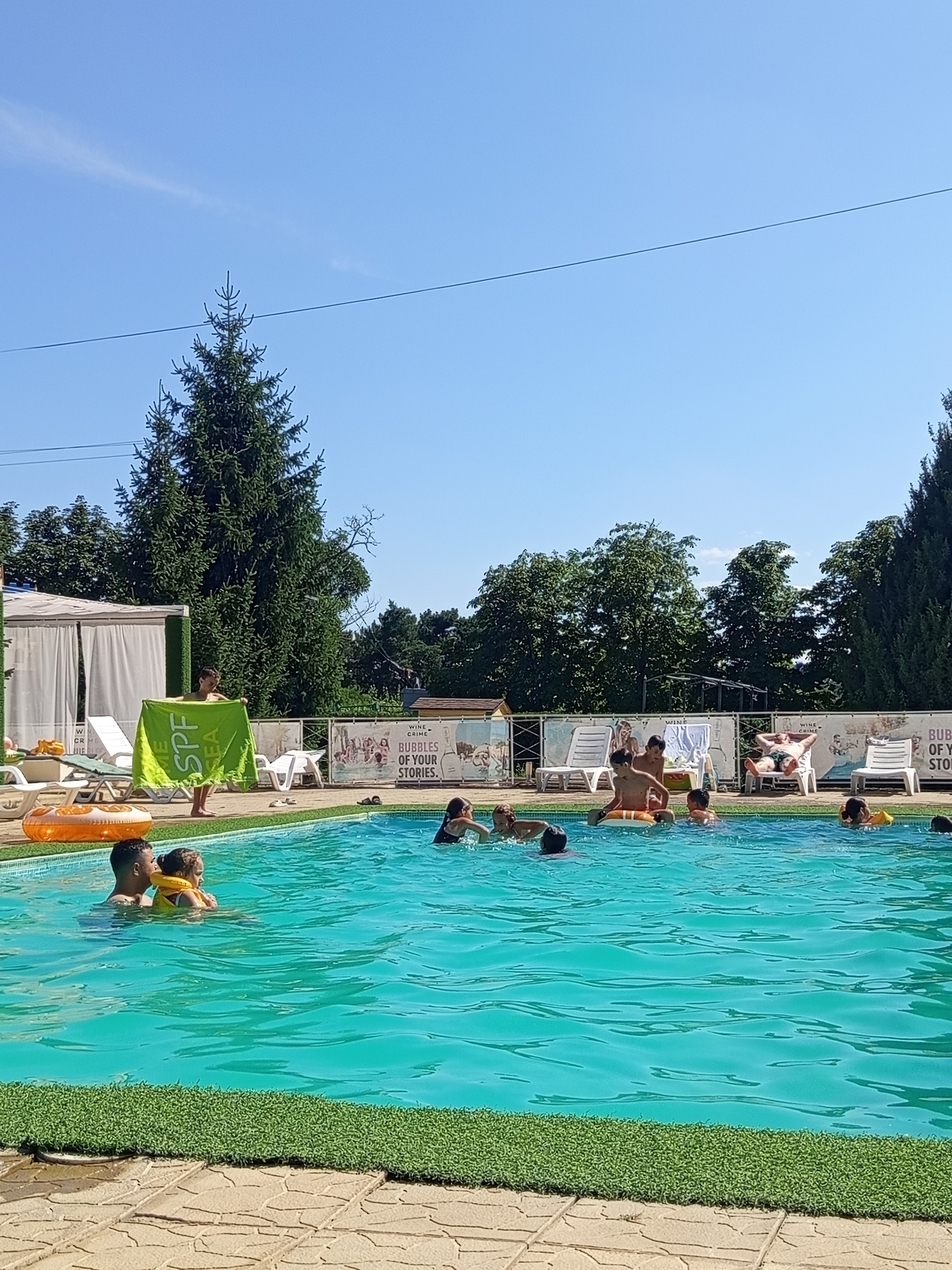 In Molovata Nouă, Moldova, a bright, sunny day is depicted at an outdoor swimming pool. The water is a vibrant turquoise, with ripples indicating activity. Several people are visible in the pool, including a man and a young child wearing a yellow flotation device, suggesting they are swimming or playing. Other individuals, including children and adults, are scattered throughout the pool, some with visible flotation devices.
Surrounding the pool is a stone patio with artificial turf lining the edge. Several white lounge chairs are arranged on the patio, with some occupied by individuals relaxing. A large orange inflatable ring is partially submerged in the water near the edge. In the background, a white tent-like structure with curtains is visible, possibly a changing room or shaded area. Trees, including a tall evergreen, form a backdrop to the scene, with a clear blue sky overhead. A banner with illustrations and text, including "WINE CRIME" and "BUBBLES OF YOUR STORIES," is mounted on a fence behind the lounge chairs, indicating a potential recreational or entertainment venue. A young person is holding a bright green towel with writing on it, possibly "SPF."