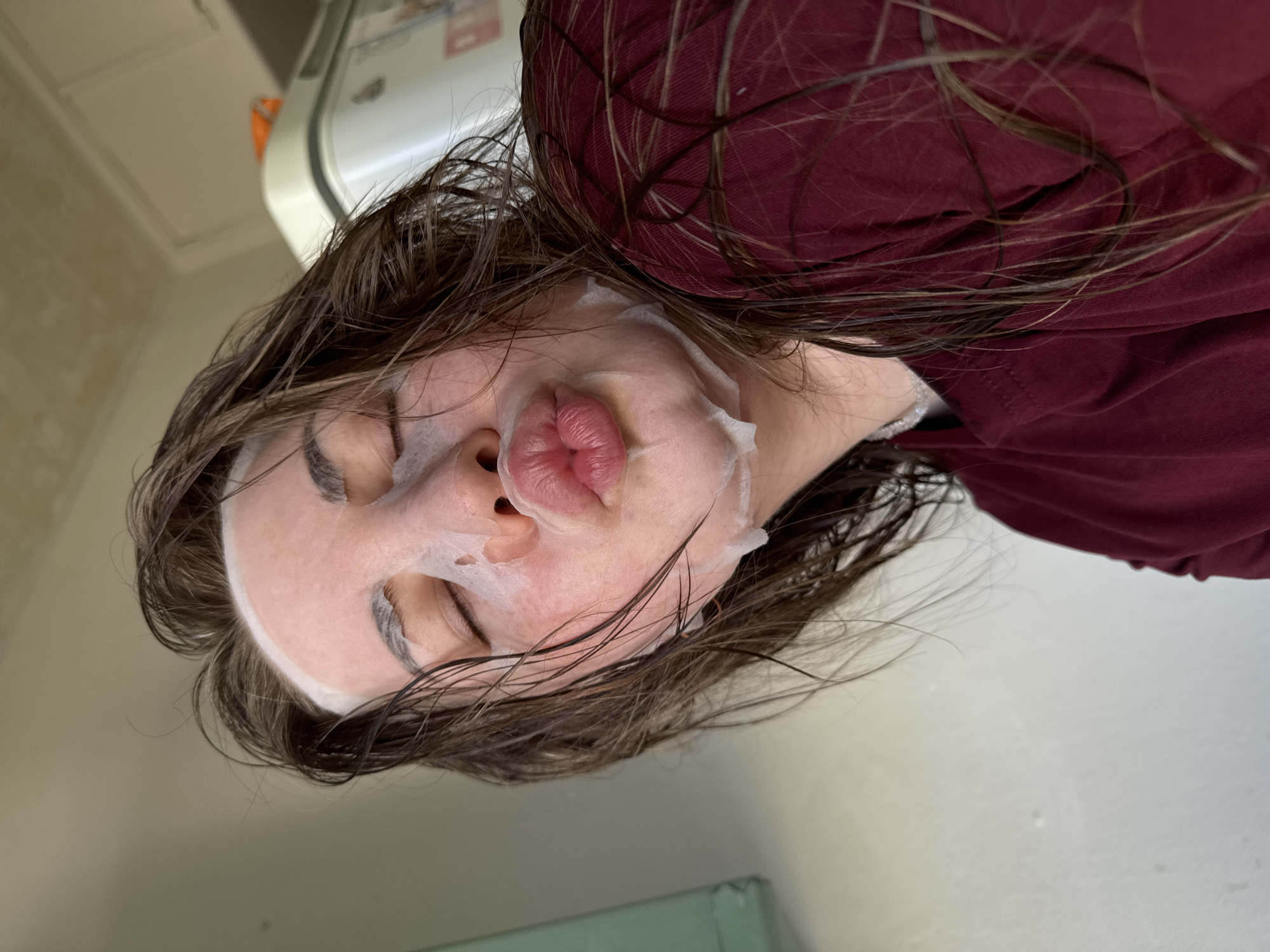 A person with wet brown hair is wearing a white sheet mask and puckering their lips. They are wearing a maroon long-sleeved shirt. The background appears to be a room with a wall and a white appliance partially visible. There is no verifiable information to determine the location.