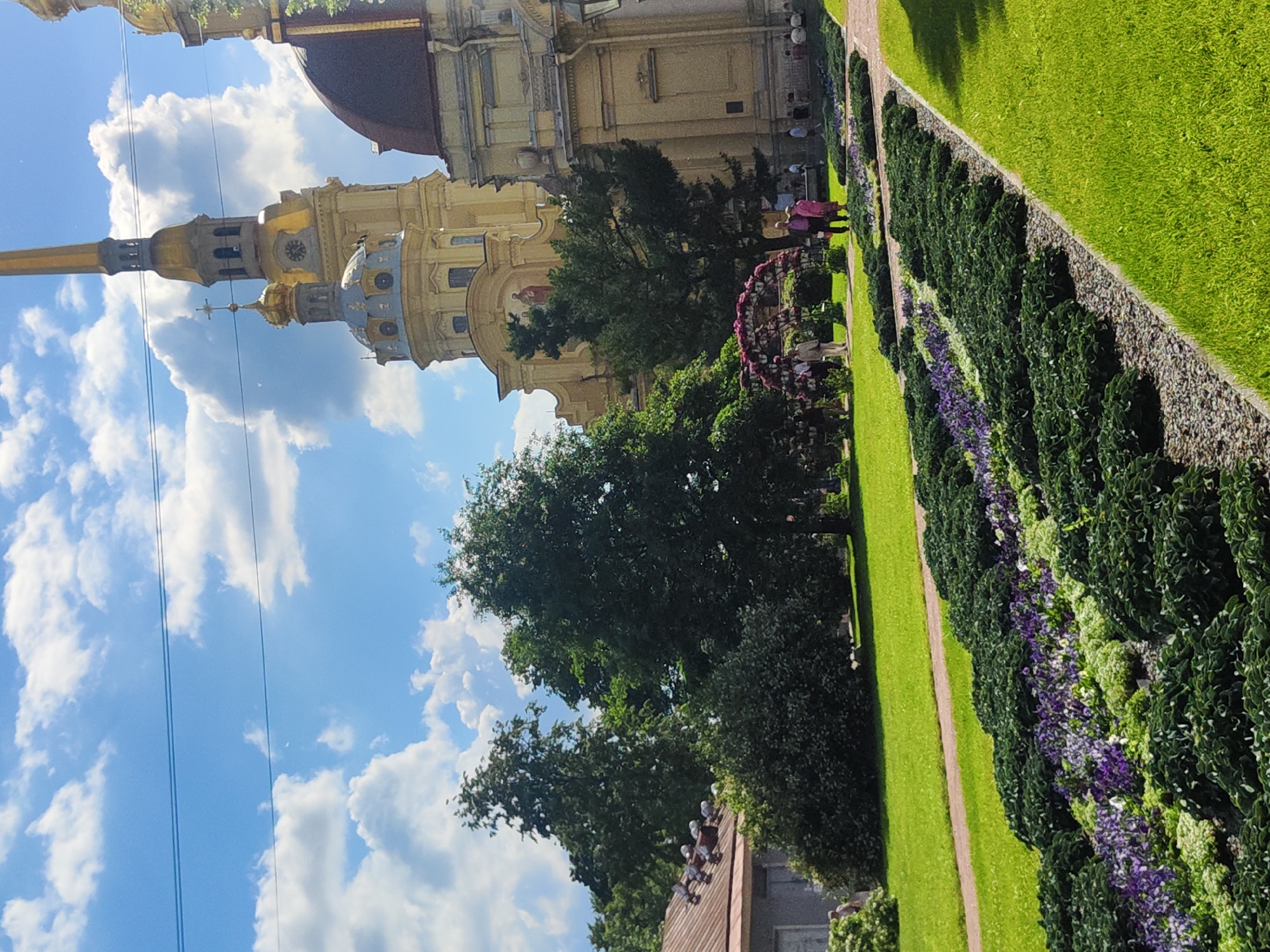 The image depicts a sunny day in the Peter and Paul Fortress in Saint Petersburg, Russia. In the background, the Peter and Paul Cathedral's spire, with its golden cross, pierces the blue sky, which is dotted with fluffy white clouds. To the right of the cathedral, a portion of another building with a red dome is visible. The middle ground features a lush green lawn and several large trees. A decorative floral archway, adorned with pink flowers, stands near a group of people gathered on the grass. In the foreground, elaborately designed flowerbeds filled with dark green foliage and purple flowers are arranged in a patterned walkway. A narrow path runs alongside the flowerbeds, leading to the green lawn.