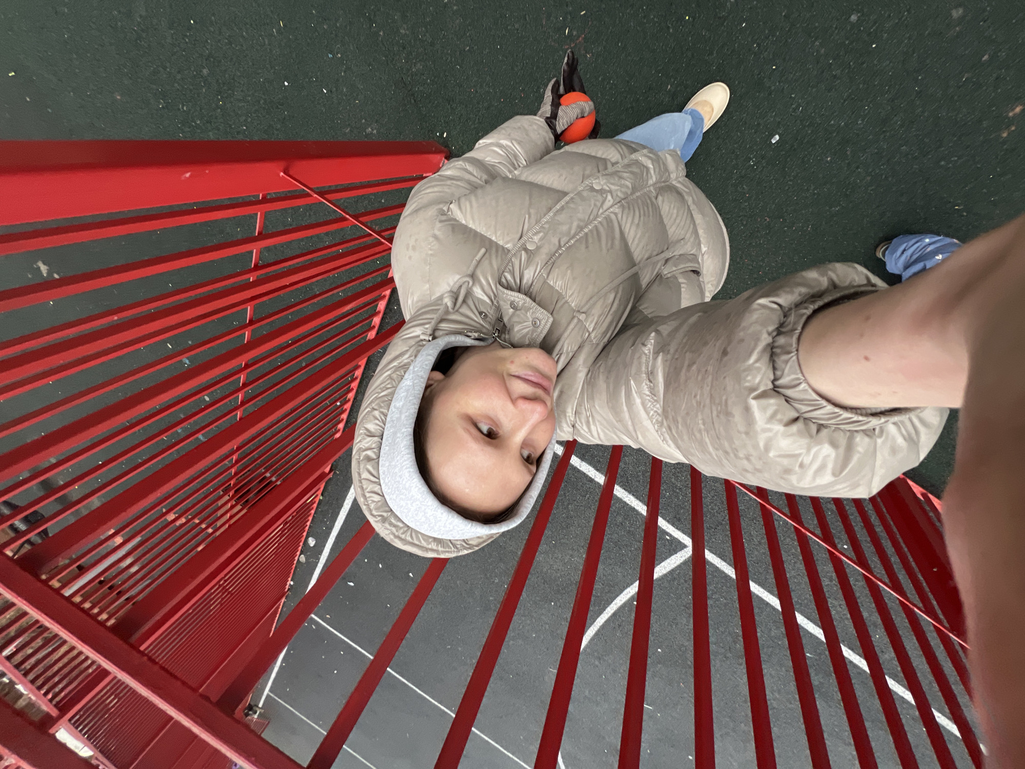 A person wearing a puffy, hooded coat is lying on a playground in Khimki, Russia. The playground has a dark green surface with white lines painted on it, and a red metal structure, likely a fence or climbing frame, is visible. The person's clothing includes blue jeans and a tan-colored shoe. An orange ball and a dark-colored glove are near the top of the frame. The person is looking up at the camera.