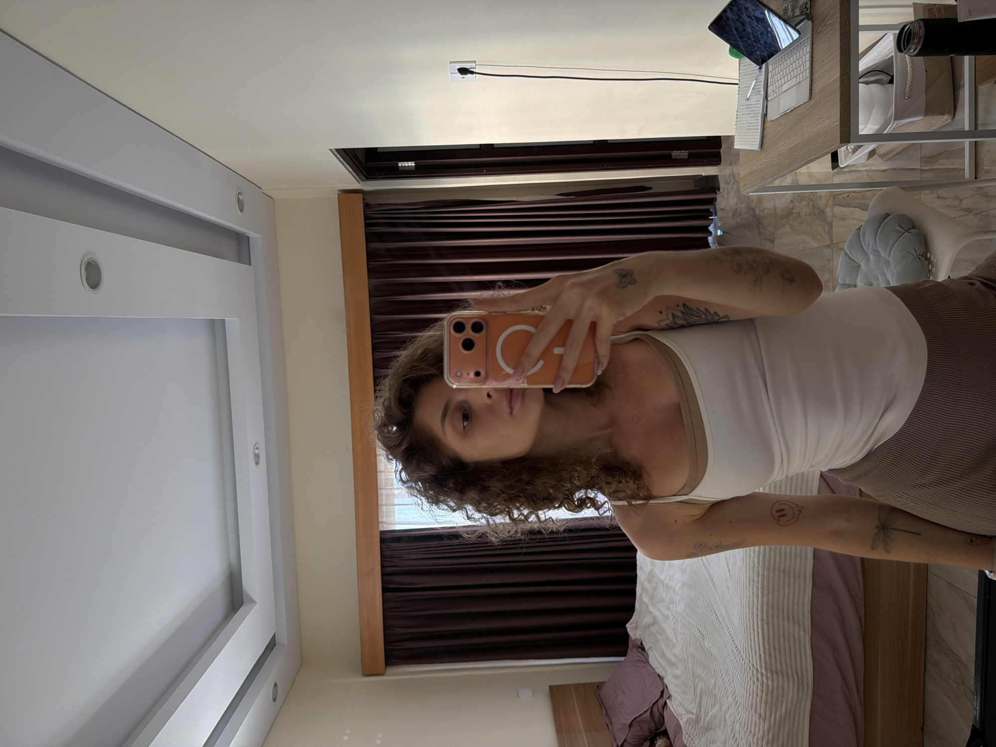 A young woman with curly brown hair takes a mirror selfie in a bedroom. She is wearing a white tank top and brown corduroy skirt. Her left arm is visible, and it has several tattoos, including a butterfly and a dragonfly. In the background, a bed with purple pillows and a white comforter is visible, as are dark brown curtains and a wooden headboard. To the right of the woman, a desk with a laptop, notebook, and pen is present. A white chair with a gray cushion is also in the room. The ceiling has a modern recessed lighting design. The woman is holding an orange smartphone in her right hand, which is reflecting the camera. The overall environment suggests a residential setting in North Kuta, Indonesia.