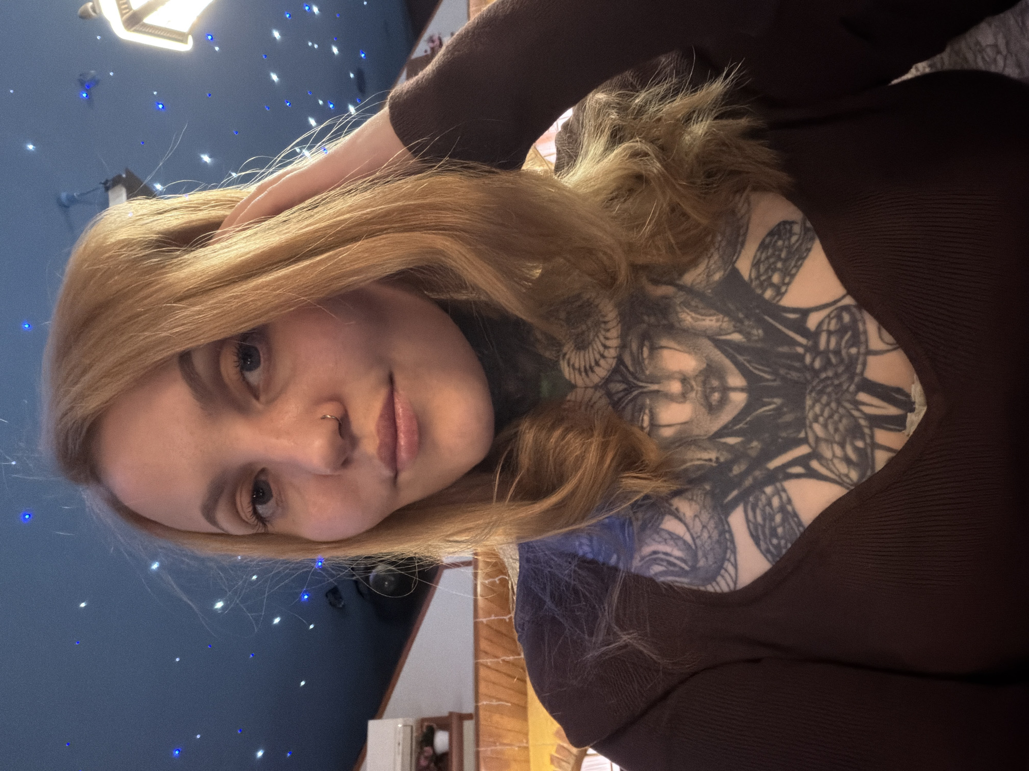 A selfie shows a fair-skinned woman with light brown hair and a nose piercing, looking at the camera. She is wearing a brown long-sleeved shirt and has a large, intricate tattoo on her upper chest and neck. Her right hand is touching her hair. The background includes a blue wall decorated with small, twinkling lights. A light fixture is visible in the upper left corner. A partial view of a wooden doorframe and a white appliance or furniture piece are also visible. There is no information to confirm that this image was taken in the city of Chaikovsky, Russia.
