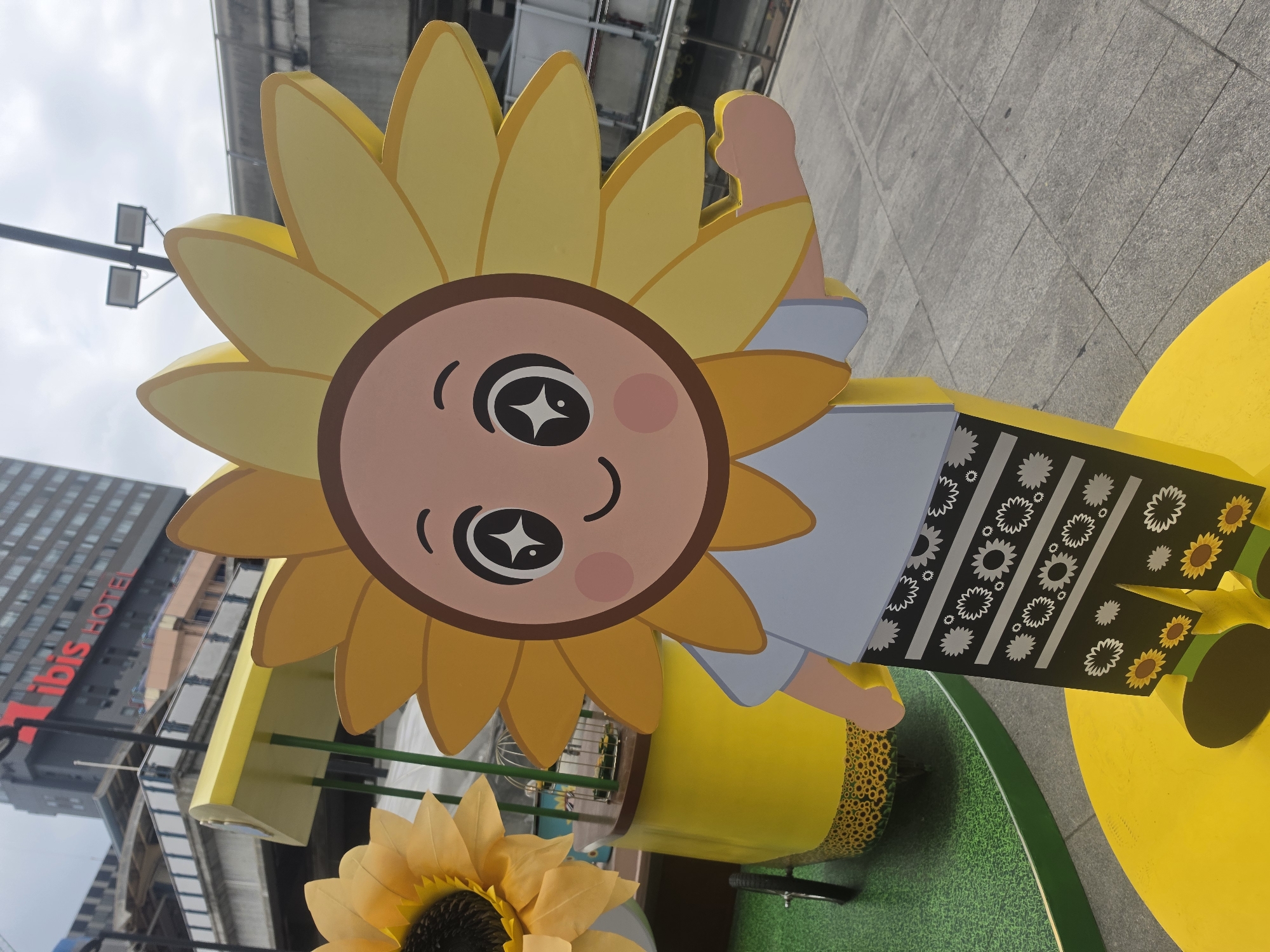 The image shows a sunny outdoor scene in Bangkok, Thailand, featuring a cartoonish sunflower figure. The figure has a round face with large, cartoonish eyes, rosy cheeks, and a smiling mouth. It is surrounded by yellow petals. The figure is wearing a light blue outfit. The background includes an Ibis Hotel building. The overall setting is an outdoor urban area with tiled pavement and artificial grass. There are also additional decorative sunflowers and other yellow structures.