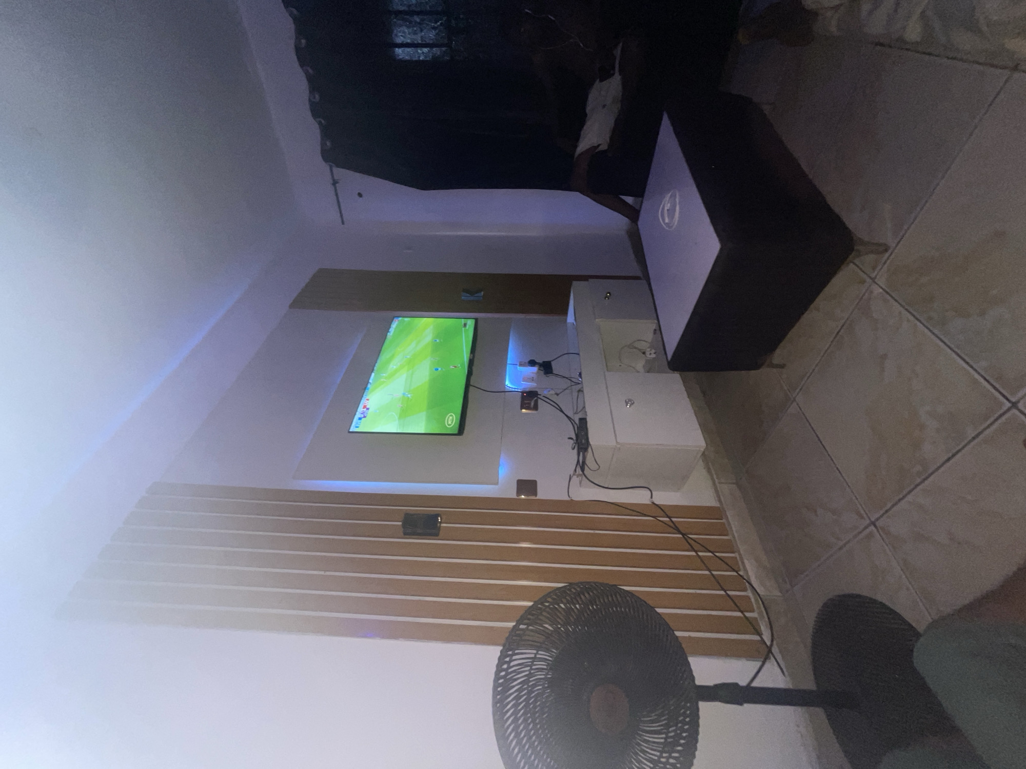 The image shows a room with a television mounted on the wall. The TV is displaying a soccer game. Below the TV is a decorative wall panel with horizontal wooden slats. A black standing fan is in the foreground. To the right of the TV is a small white cabinet and a black coffee table. The floor is tiled. Someone is standing to the left of the coffee table. The location is likely a residence in Lokoja, Nigeria.