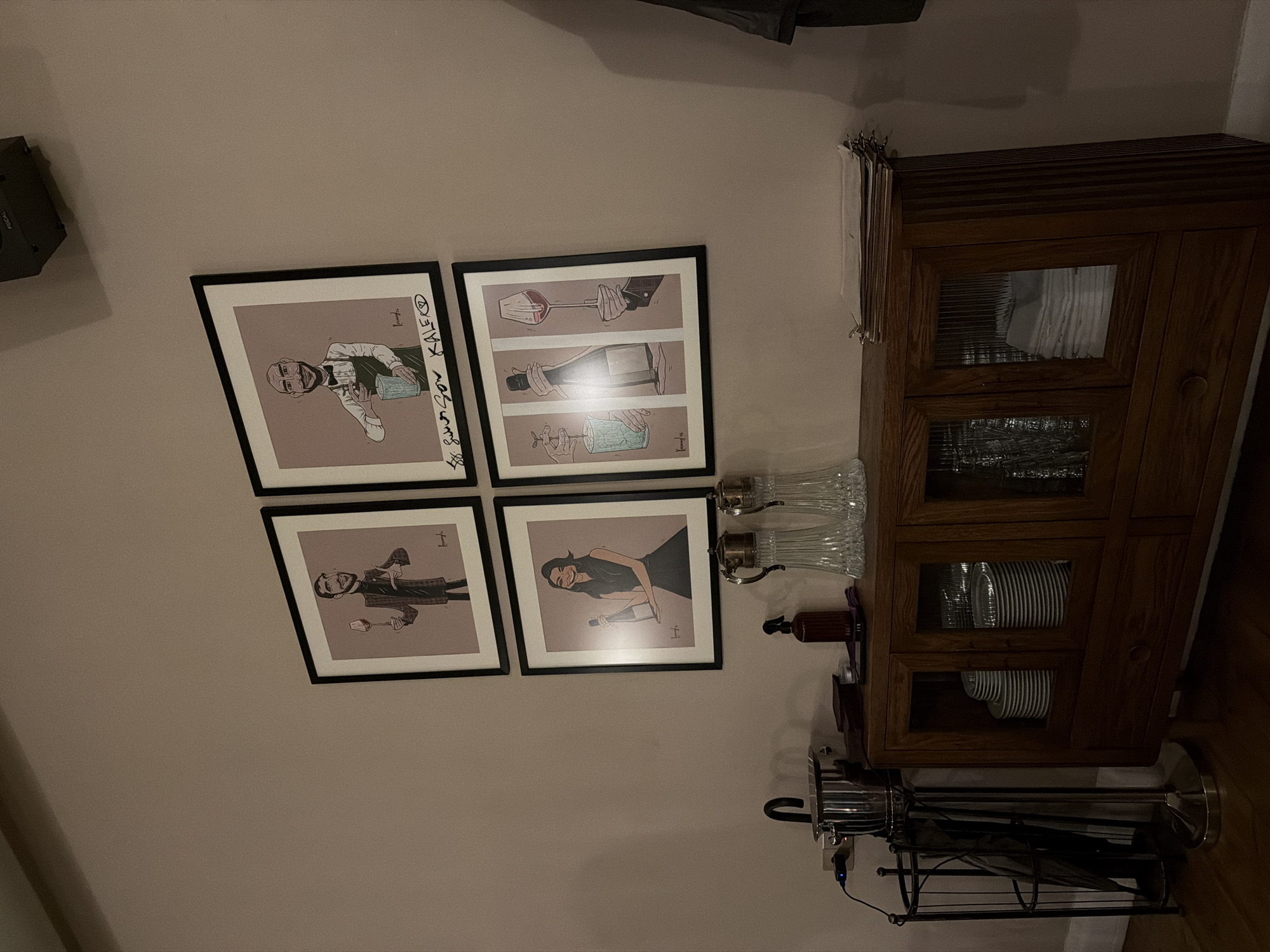 The image depicts an interior wall with four framed art prints arranged in a square formation. To the right of the prints is a wooden cabinet with glass-paneled doors, behind which plates and glasses are visible. On top of the cabinet are several glass bottles and a small bottle of alcohol. Next to the cabinet is an umbrella stand. The environment appears to be inside a house or apartment. It is not possible to determine the specific location in Yerevan, Armenia from the image alone.