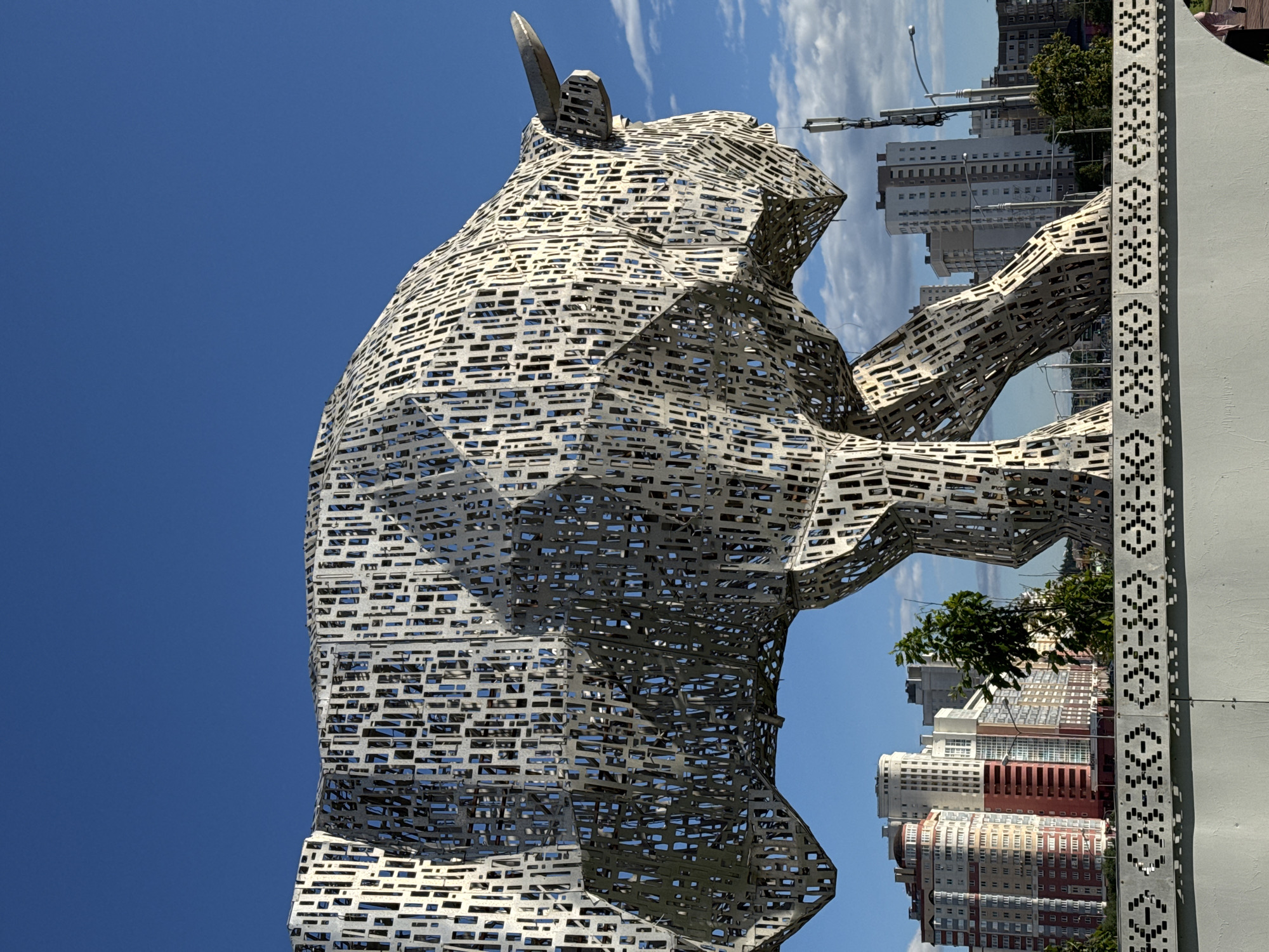 The image shows a large, abstract sculpture of a bull made of metal with a geometric, perforated design. The sculpture is positioned in the foreground, with its head and front legs visible. In the background, a cityscape of Minsk, Belarus, is visible, featuring numerous tall apartment buildings. The sky is blue with some scattered clouds. The sculpture is situated on a concrete base with a decorative border. A street lamp and a communication tower are also present in the background.
