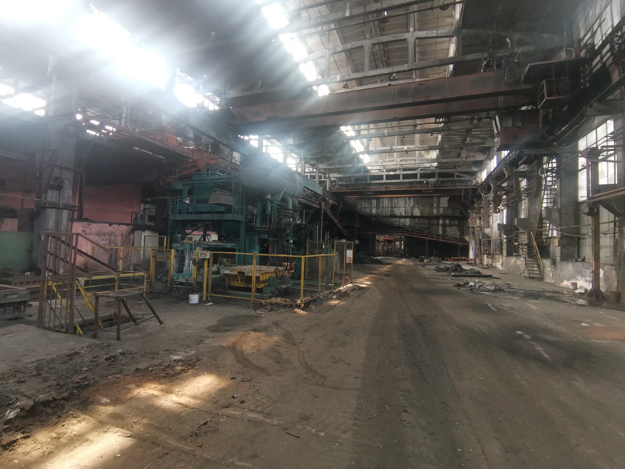 The image depicts the interior of an abandoned factory in Zhivkovo, Bulgaria. The factory appears to be a large, industrial space with high ceilings and exposed metal beams. Sunlight streams through the openings in the roof, illuminating dust and debris scattered across the dirt floor. In the foreground, there are various pieces of industrial machinery surrounded by yellow safety railings. Towards the back of the space is a corridor filled with debris. The overall environment suggests a state of disuse and disrepair. No people are visible.