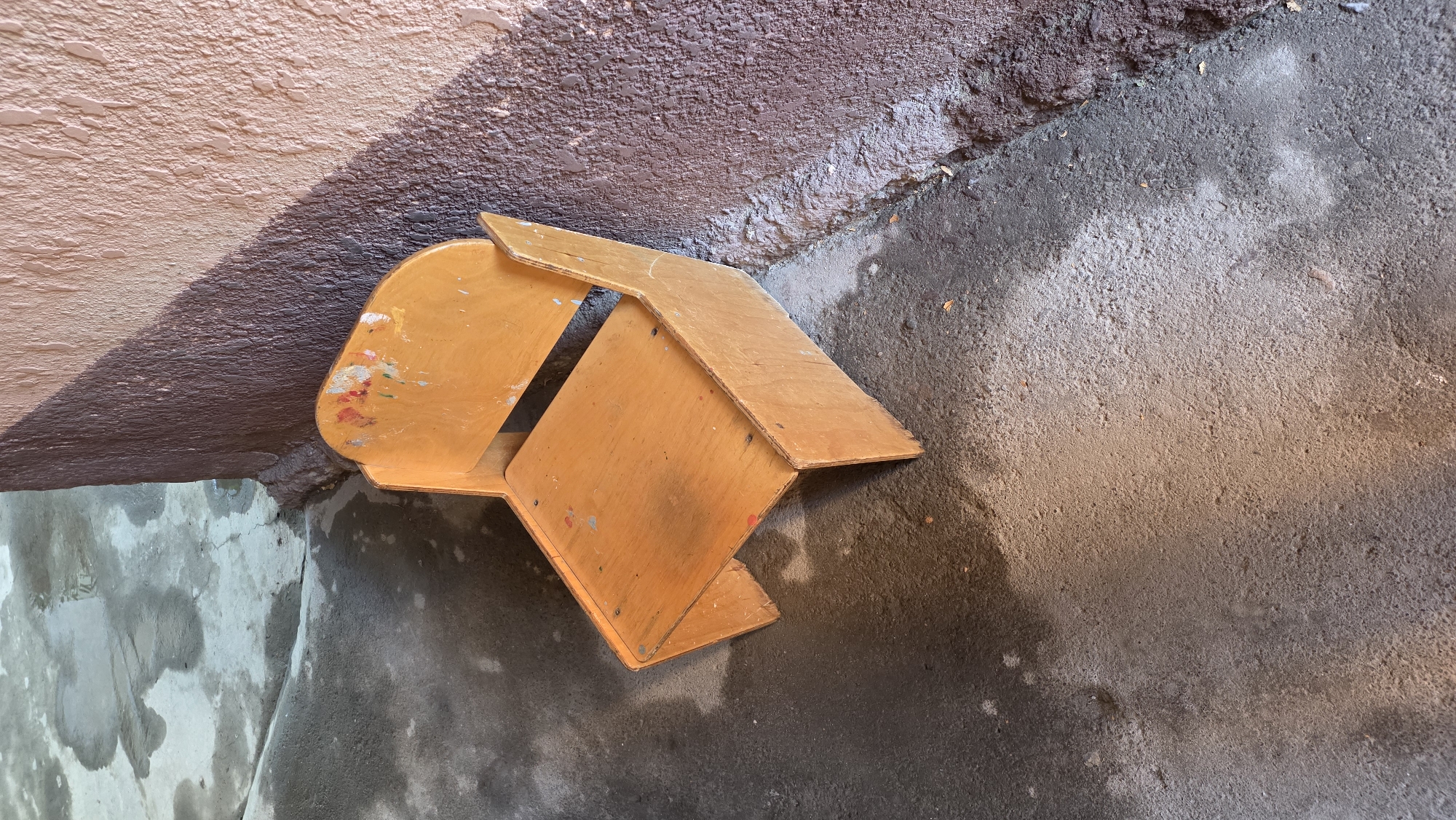 An outdoor scene in Tokmok City, Kyrgyzstan, shows a small, light-brown wooden chair situated on a damp, textured concrete ground. The chair has a simple design with a seat and a backrest, and appears to be used, with some colorful paint splatters visible on its surface. To the right of the chair, a rough, textured wall in shades of brown and pink rises, casting a distinct shadow across the ground. The ground itself shows signs of moisture and is uneven, with areas of darker wet patches and lighter dry sections. The overall impression is one of an aged, possibly abandoned or neglected, outdoor space.