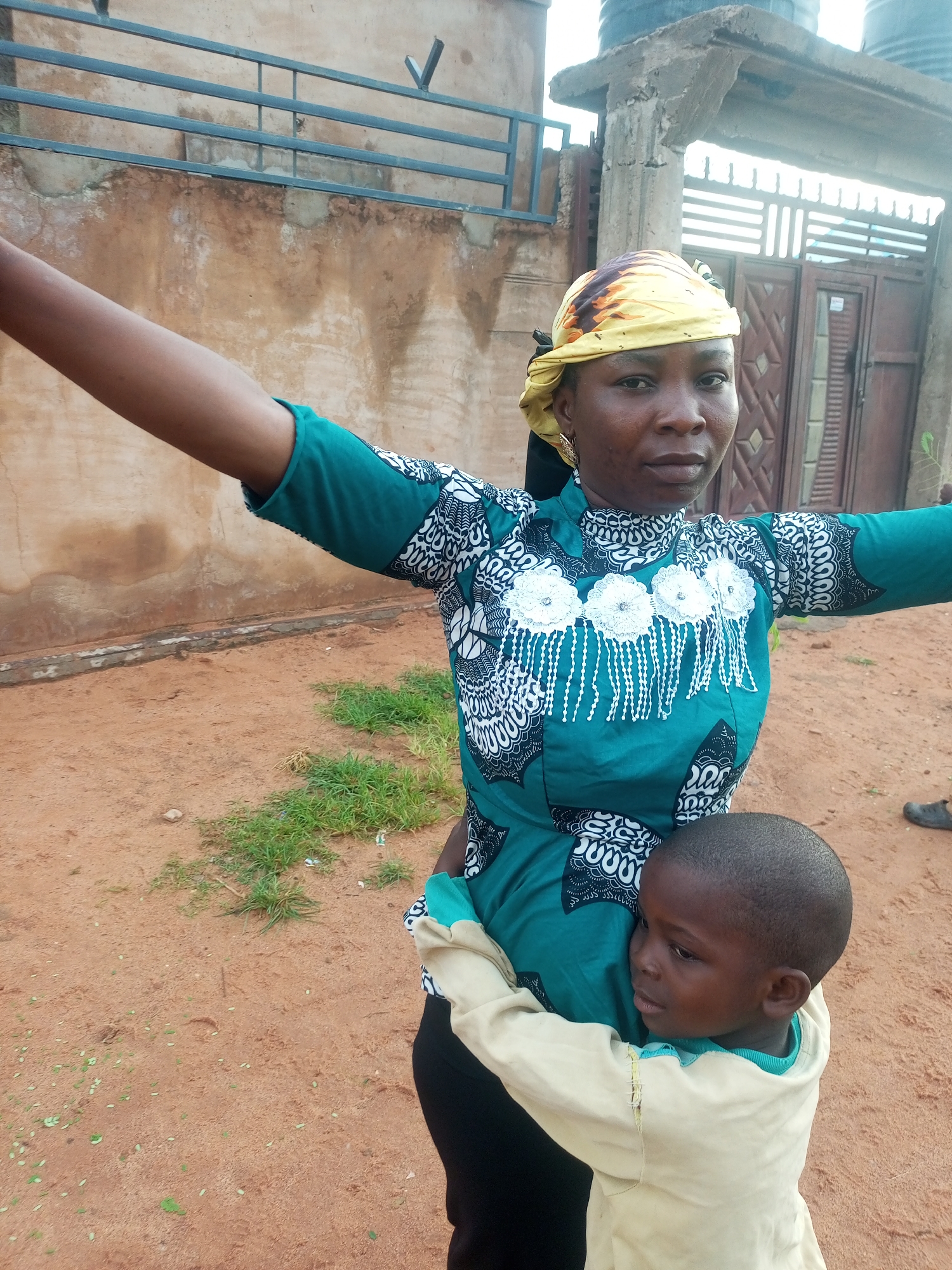 A medium shot shows a woman and a young child in what appears to be a residential area in Tempure, Nigeria. The woman, who is wearing a teal and white patterned long-sleeved dress and a yellow headscarf, is standing with her arms outstretched, as if embracing the child or posing for the photo. The child, a boy dressed in a cream-colored long-sleeved shirt, is hugging the woman from the front, with his head resting on her side.
The background consists of a rustic building with a light brown, textured wall, possibly made of mud or stucco. A metal railing is affixed to the wall. To the right of the building, there is a partial view of an entryway with a metal gate and two water tanks on top of a concrete structure. The ground is covered in reddish-brown dirt, with patches of green grass scattered about. The lighting suggests it is daytime.