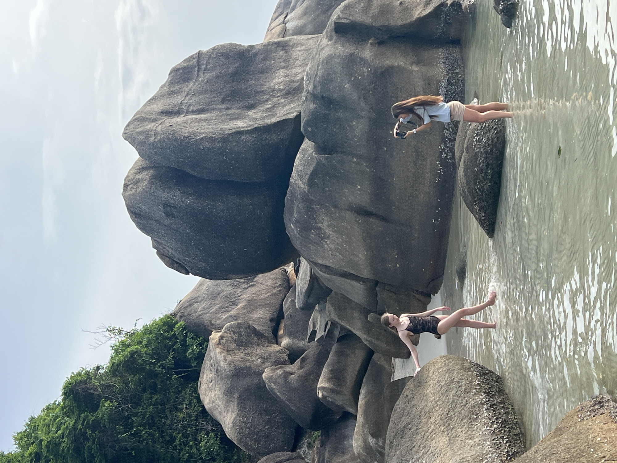 Two young women are standing in shallow ocean water near a rocky coastline. The woman on the left, wearing a black one-piece swimsuit, is positioned with her left hand on a large boulder, her right leg slightly forward. The woman on the right, wearing a light blue shirt and tan shorts, is holding a camera and appears to be taking a photo of the woman on the left. Behind them are several large, grey, weathered boulders, some covered in barnacles, and lush green vegetation grows on the rocks in the background. The water is clear and calm, with ripples reflecting the sky. The location is Baan Chaweng Noi, Thailand.