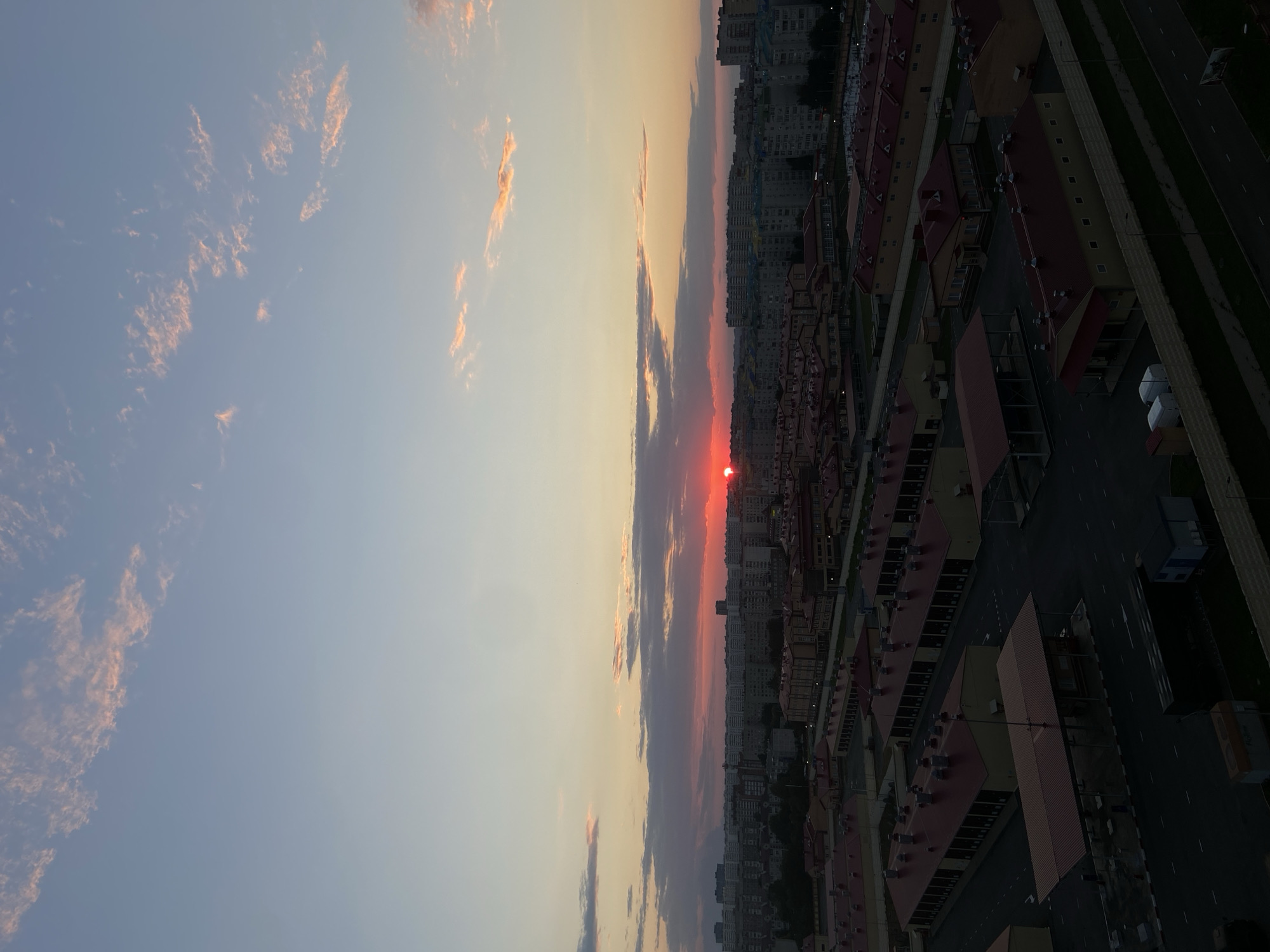 This is a vertical, eye-level, outdoor shot of a cityscape at sunset. The sun is a bright red orb, partially obscured by the horizon line, casting a warm glow across the sky. The sky is a gradient of soft blues, oranges, and pinks, with wispy clouds catching the last rays of sunlight. In the foreground, the roofs of buildings are visible, mostly in shades of red and yellow. The buildings appear to be residential or commercial, with a uniform architectural style. Streets and sidewalks weave between the buildings, and a few parked vehicles are visible. In the background, taller apartment buildings rise above the lower structures, indicating a densely populated urban area. The overall mood is peaceful and serene, capturing the beauty of a sunset over a city. The image appears to be taken from a high vantage point, offering a panoramic view of the urban landscape. The time of day is clearly sunset, indicated by the sun's position and the colors in the sky. There are no people visible in the frame. The location is identified as Krasnodar, Russia.