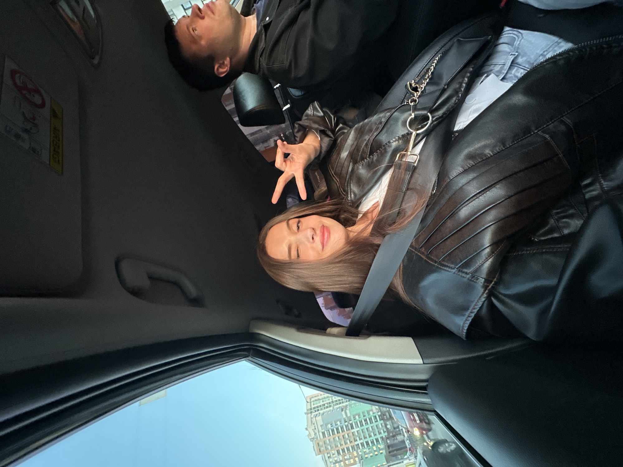 The image captures a playful moment inside a car, rotated counter-clockwise. In the foreground, a young woman with long brown hair is seated, wearing a dark brown or black leather-look jacket over a white top, secured by a seatbelt. She smiles towards the camera, making a "peace" or "V" sign with her right hand. A small bag with a gold chain strap and a keyring is visible near her jacket. Behind her, a man is partially visible, seated in another car seat, wearing a dark jacket/shirt, looking forward.
The car's interior features a dark headliner, a grab handle, and an "AIRBAG" warning label on the passenger-side sun visor, accompanied by other symbols and text. Through the car window, a bright blue sky suggests clear daytime weather. An urban setting is visible outside, with modern buildings featuring grid-like structures and some green elements. The scene depicts travel through a city, consistent with the location context of Yekaterinburg, Russia. The woman's expression conveys happiness and cheerfulness.