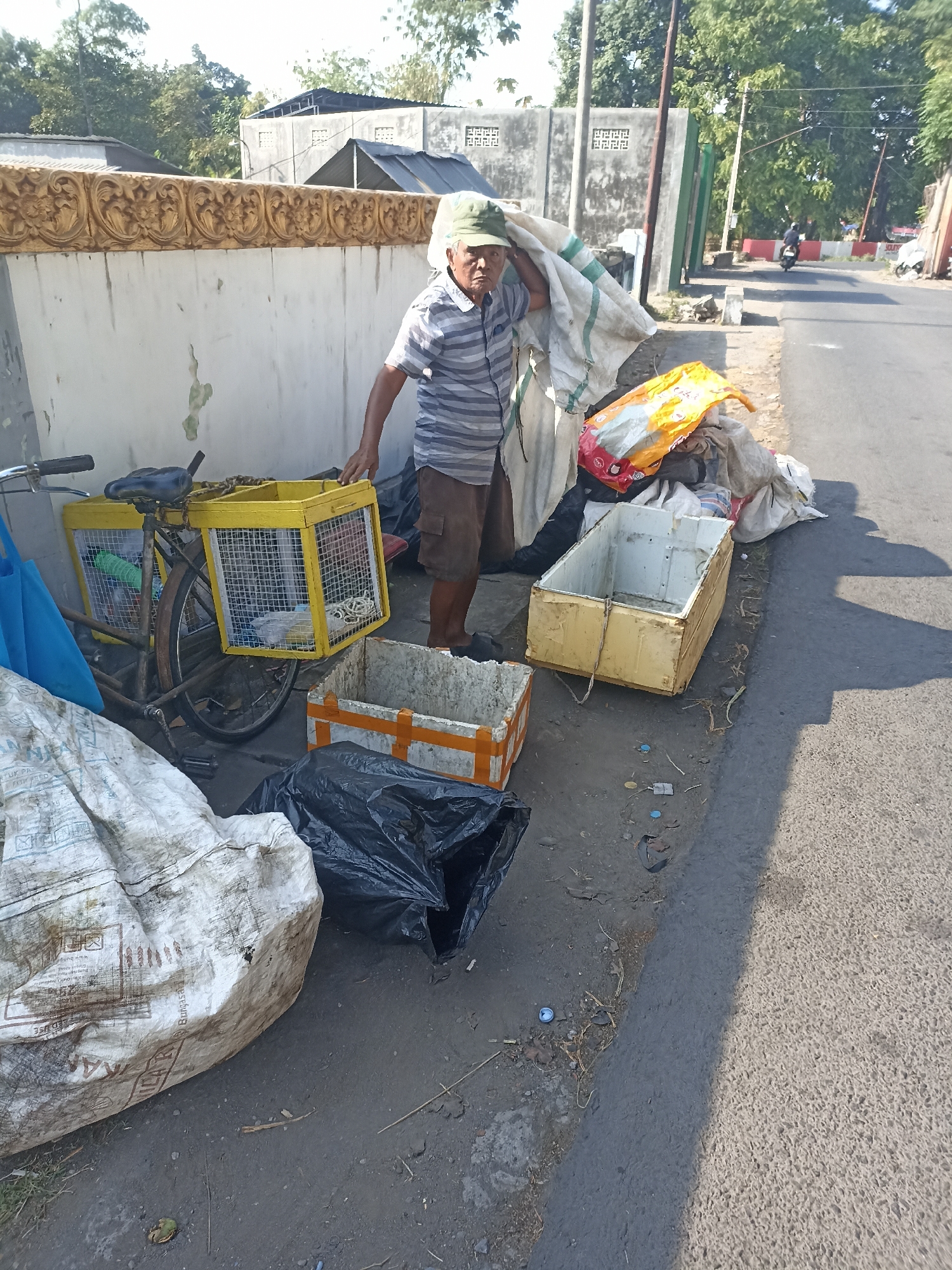 The image depicts a man in Mojopanggung, Indonesia, standing next to a bicycle laden with yellow cages and a large sack of what appears to be recyclable materials. The man is wearing a striped shirt, shorts, and a green cap, and is holding the sack over his shoulder. In the foreground, there are several discarded items, including plastic bags, a white styrofoam box, and a smaller orange-and-white taped container. The scene is set on the side of a paved road, with a wall and trees in the background. A motorcycle is visible in the distance on the road. The overall impression is of a person engaged in collecting or transporting waste materials.