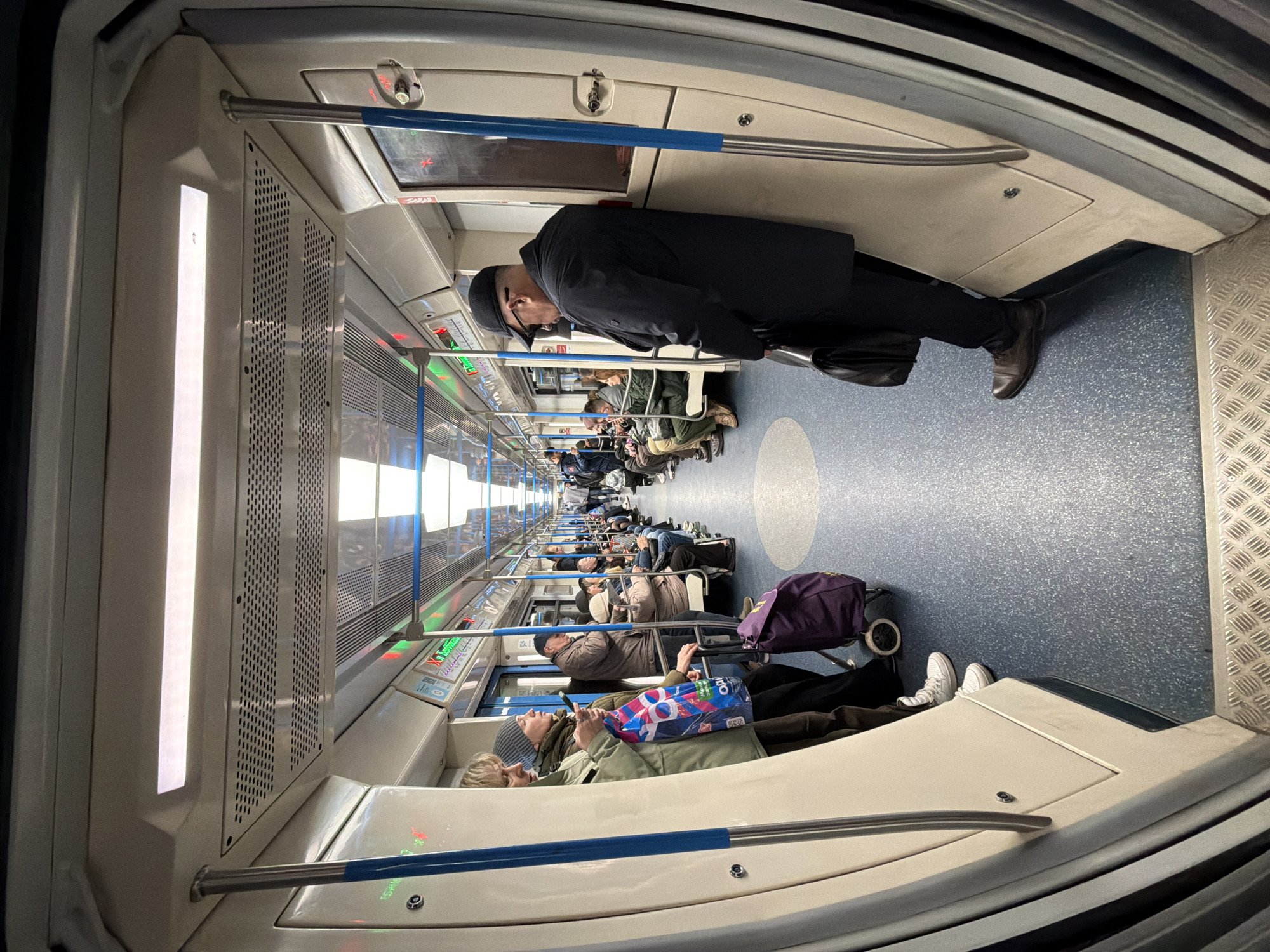 Here is a forensic description of the image:
**Scene:** The image depicts the interior of a subway train in Moscow, Russia. It is populated with numerous passengers.
**Environment:** The subway car features white walls, ceiling, and seating. A blue horizontal bar runs along the sides of the car. The floor is predominantly blue with a circular pattern and diamond-shaped metallic tread.
**People:** There are numerous passengers visible, mostly seated. One man wearing a black coat, hat, and glasses is standing. Passengers appear to be of varying ages and are dressed in casual clothing. Some passengers have luggage.
**Objects:** Luggage includes a purple suitcase with wheels and a patterned shopping bag.
**Actions:** The passengers are seated, likely commuting. The standing man is facing towards the seated passengers.
**Interactions:** There are no immediately obvious interactions among the passengers. The focus of most individuals seems to be self-directed.