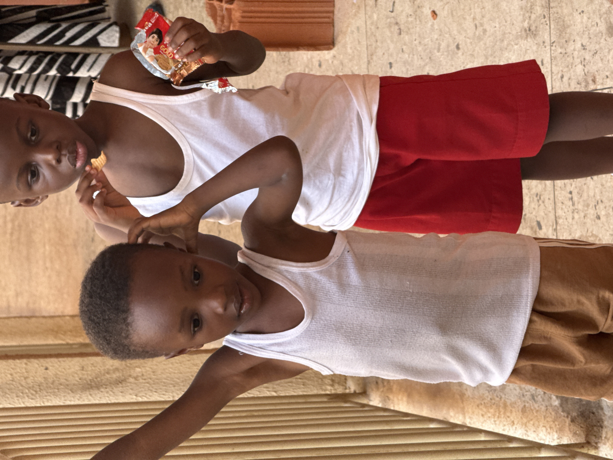 Two young African boys are depicted indoors. One boy is eating a cracker from a package, wearing a white tank top and red shorts. The other boy is wearing a white tank top and brown pants. The image's background suggests an interior setting, potentially in Abuja, Nigeria.